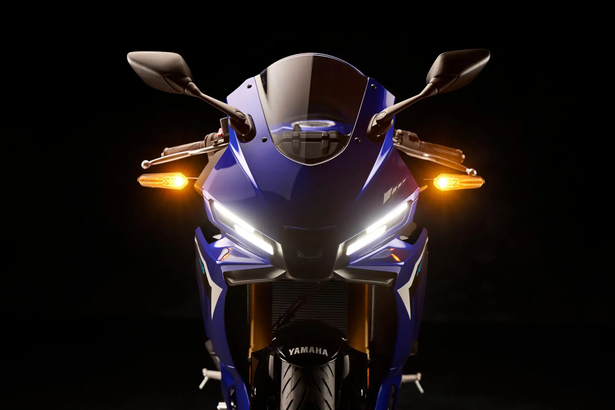 2025-Yamaha-YZF-R320-EU-Detail-006-03 Vorderansicht einer blauen Yamaha YZF-R320 mit eingeschalteten Scheinwerfern und Blinkern.