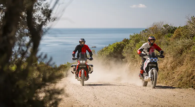 Zwei Personen fahren auf Ducati DesertX Motorrädern auf einer staubigen Küstenstraße.