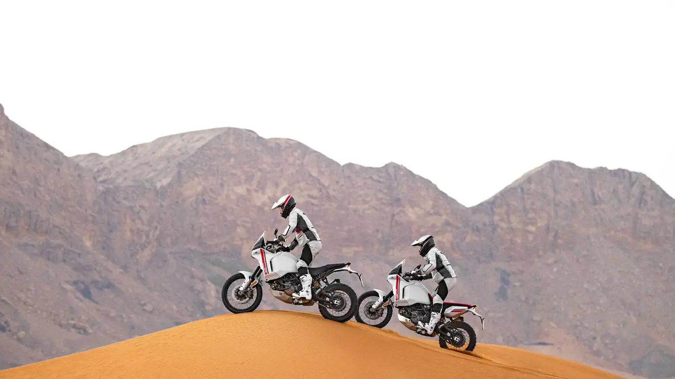 Zwei Personen fahren auf Ducati DesertX Motorrädern über eine Sanddüne.
