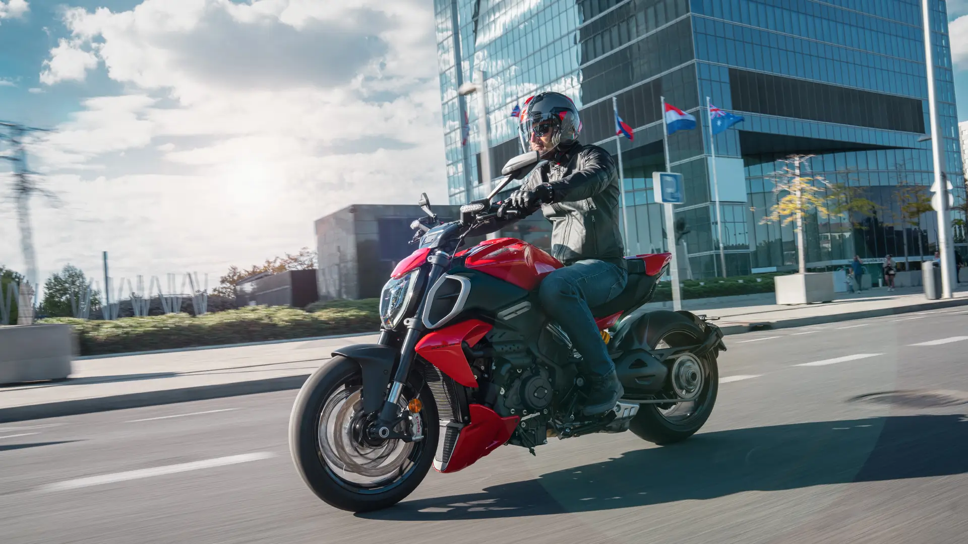 Ein Motorradfahrer fährt eine rote Ducati Diavel V4 vor einem modernen Bürogebäude.