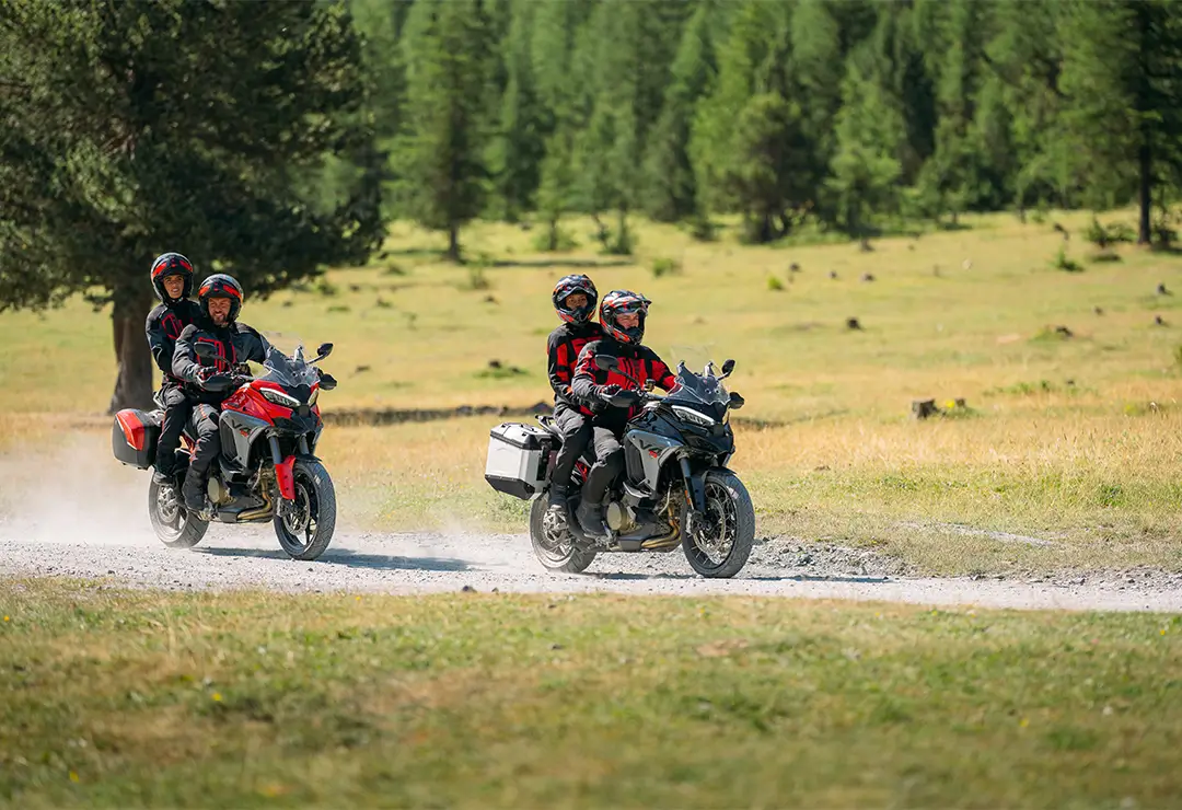 Zwei Ducati Multistrada V4 Motorräder mit Fahrern und Beifahrern auf einem Schotterweg in einer grünen Landschaft.