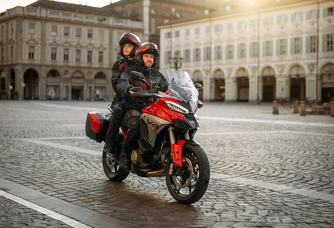 Ducati-MY25-Multistrada-V4-motore-carousel-imgtext-full-1080x740-03 Zwei Personen fahren auf einer Ducati Multistrada V4 durch einen leeren Platz.