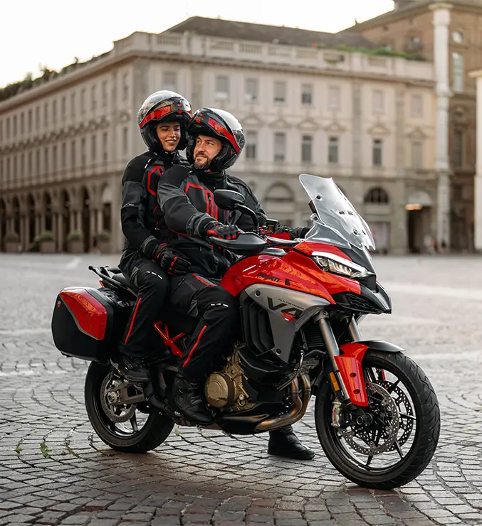 Ducati-MY25-Multistrada-V4-overview-carousel-imgtext-677x740 Zwei Personen in Motorradkleidung sitzen auf einer Ducati Multistrada V4 auf einem gepflasterten Platz.