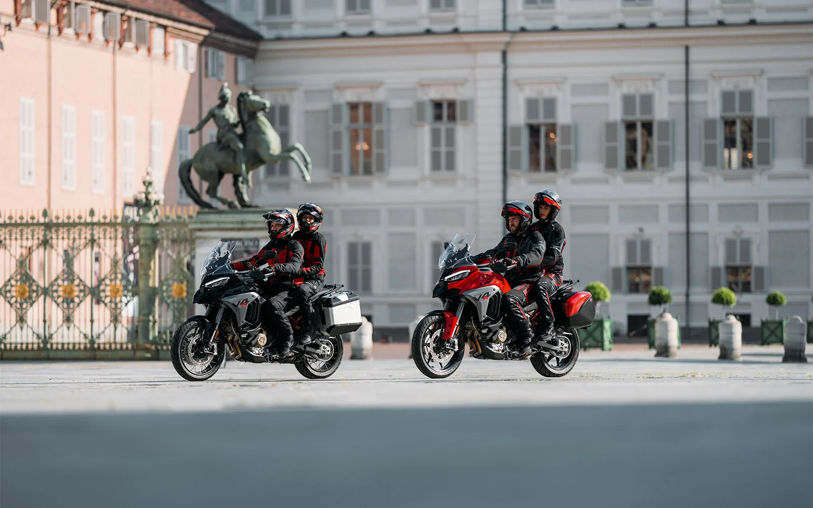 Zwei Ducati Multistrada V4 Motorräder mit Fahrern und Beifahrern vor einem historischen Gebäude.