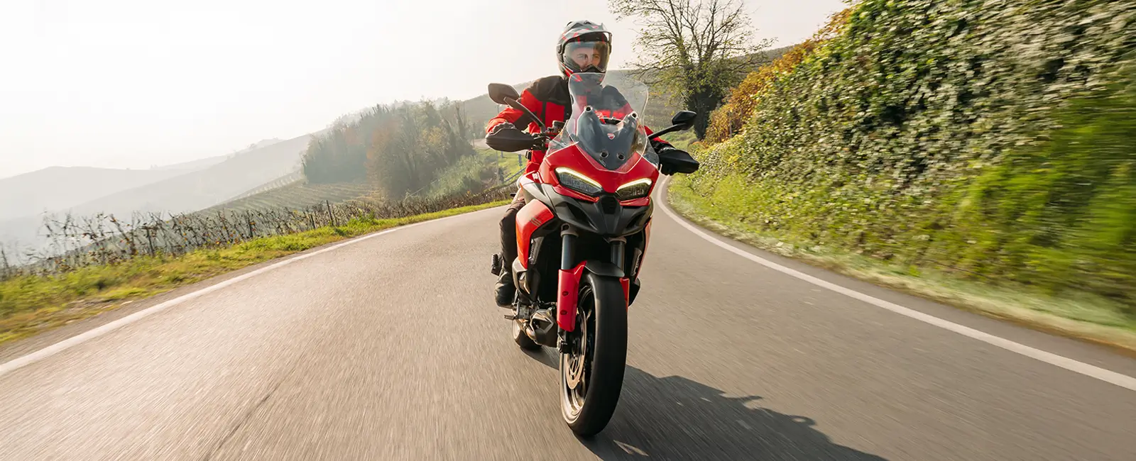 Ducati-Multistrada-V2-DWP25-overview-carousel-hero-link-1600x650-03 Ein Motorradfahrer fährt eine rote Ducati Multistrada V2 auf einer kurvigen Landstraße.
