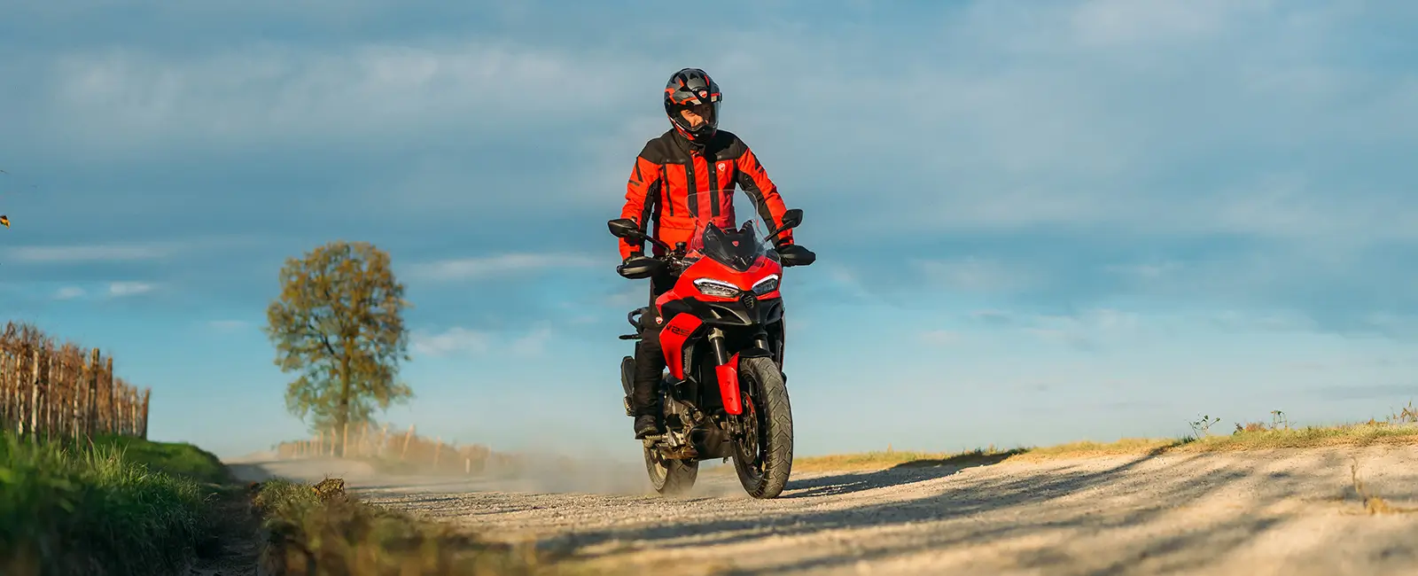 Ducati-Multistrada-V2-DWP25-overview-carousel-hero-link-1600x650-05 Ein Motorradfahrer auf einer Ducati Multistrada V2 fährt auf einer ländlichen Straße.