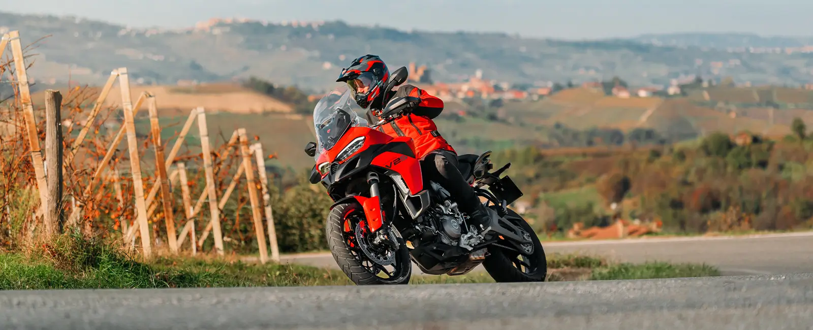 Ducati-Multistrada-V2-DWP25-overview-carousel-hero-link-1600x650-06 Ein Motorradfahrer fährt eine rote Ducati Multistrada V2 durch eine ländliche Landschaft.
