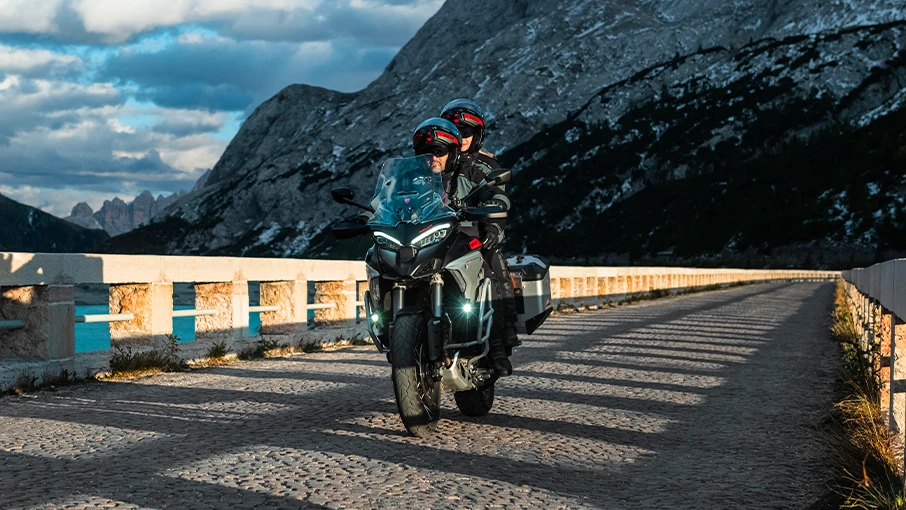 Zwei Personen fahren auf einer Ducati Multistrada V4 Rally über eine malerische Brücke in den Bergen.
