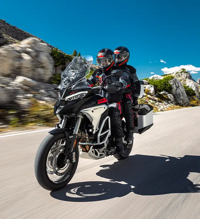 Zwei Personen fahren auf einer Ducati Multistrada V4 Rally auf einer kurvigen Bergstraße.
