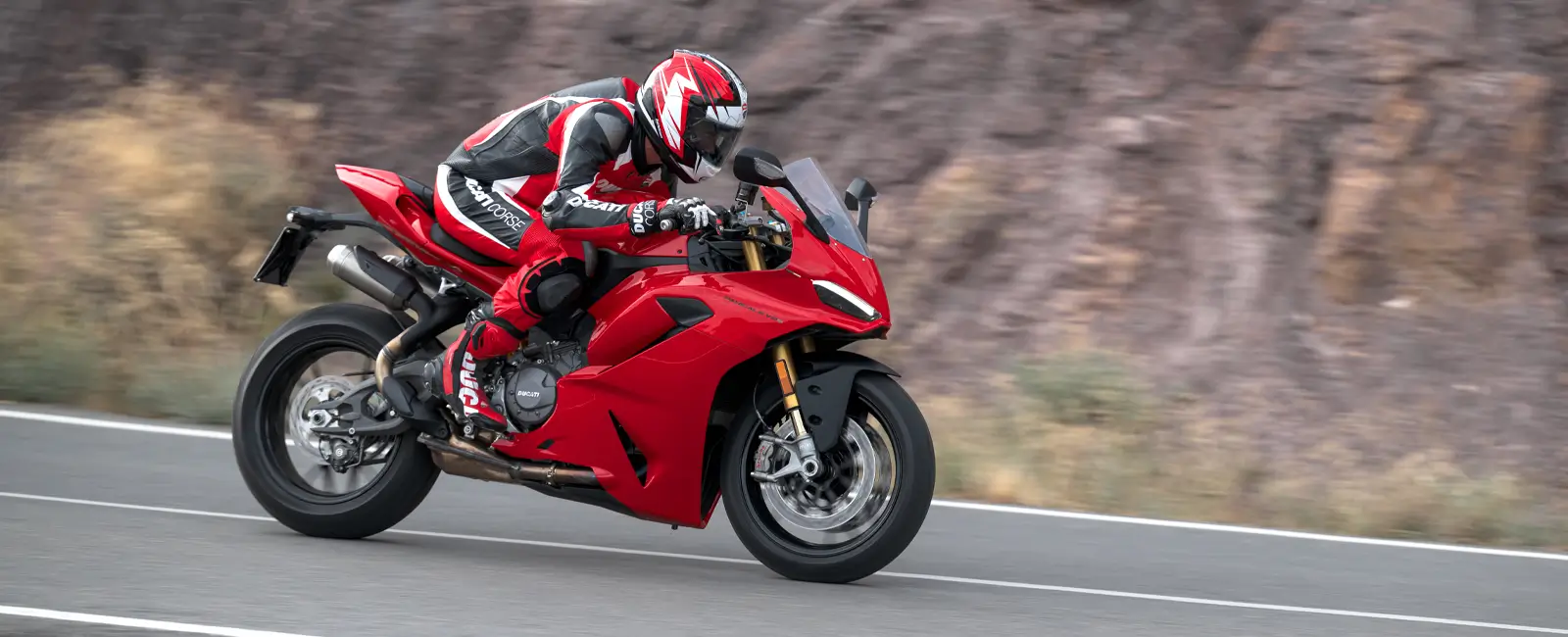 Ducati-Panigale-V2-V2S-Overview-carousel-hero-link-1600x650-03 Ein Motorradfahrer auf einer roten Ducati Panigale V2 fährt auf einer Straße.