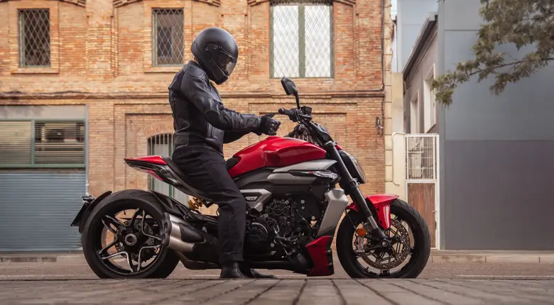 Ducati-XDiavel-V4-overview-mosaic-gallery-780x430-04 Ein Motorradfahrer sitzt auf einer Ducati XDiavel V4 vor einem Backsteingebäude.