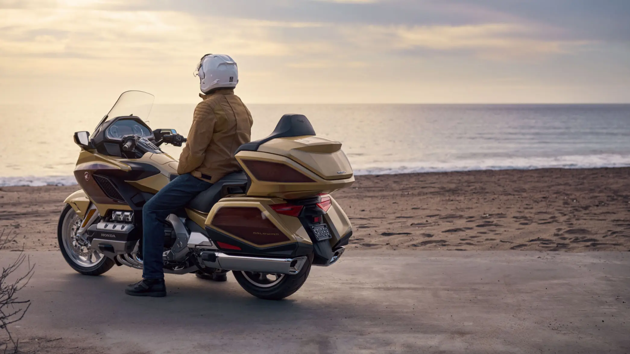 Eine Person sitzt auf einem goldenen Honda GL1800 GOLD WING TOUR Motorrad am Strand und blickt aufs Meer.