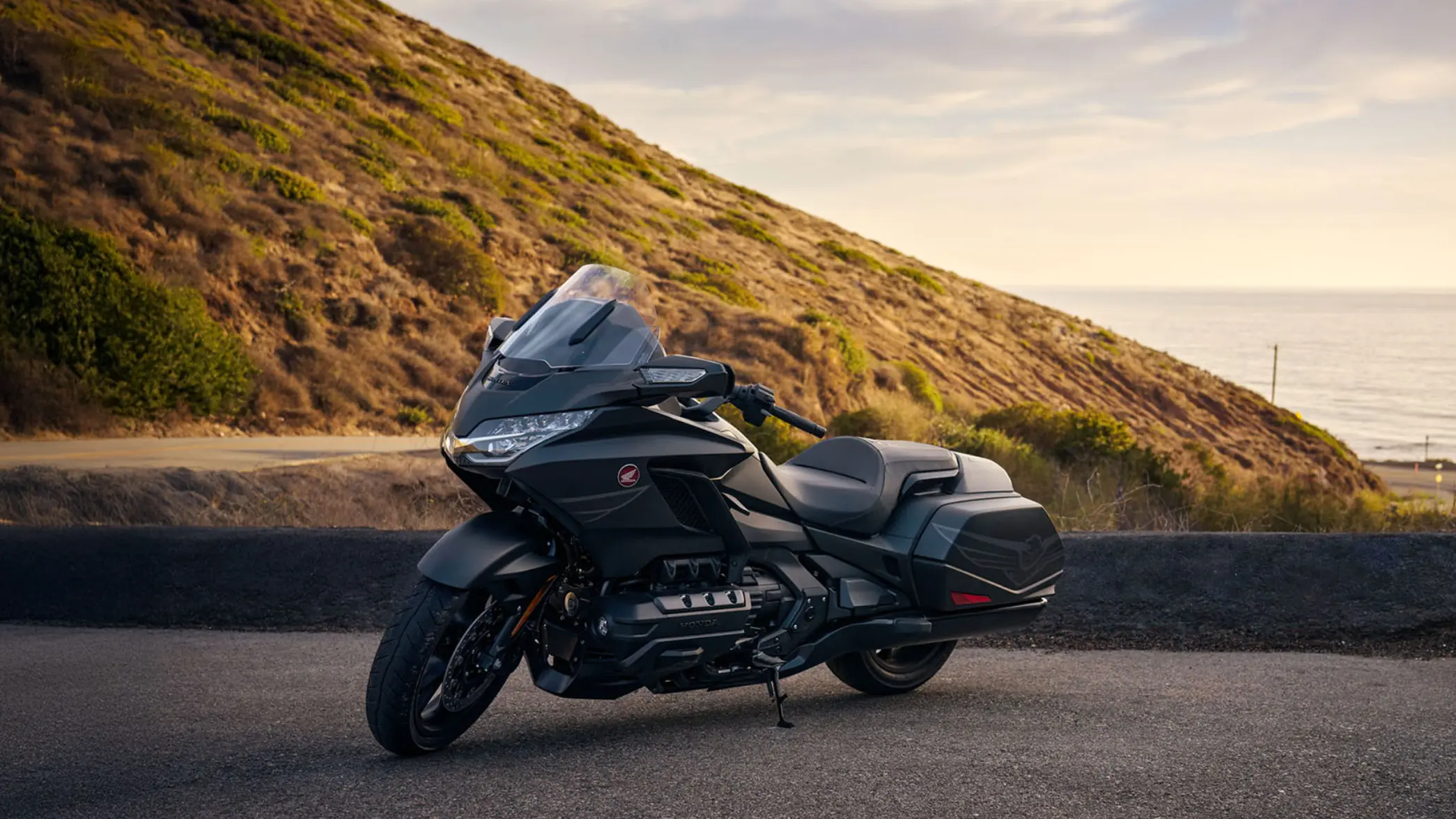fb_r_w Schwarzes Honda-Motorrad, geparkt auf einer Straße vor einem Hügel mit Meerblick im Hintergrund.
