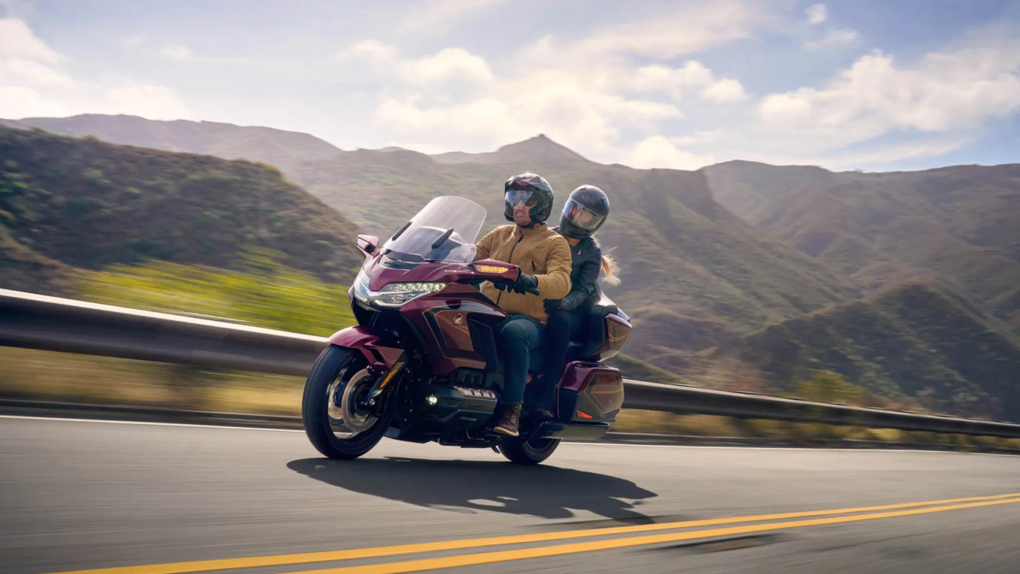 Zwei Personen fahren auf einem Honda GL1800 GOLD WING TOUR auf einer Straße durch eine bergige Landschaft.