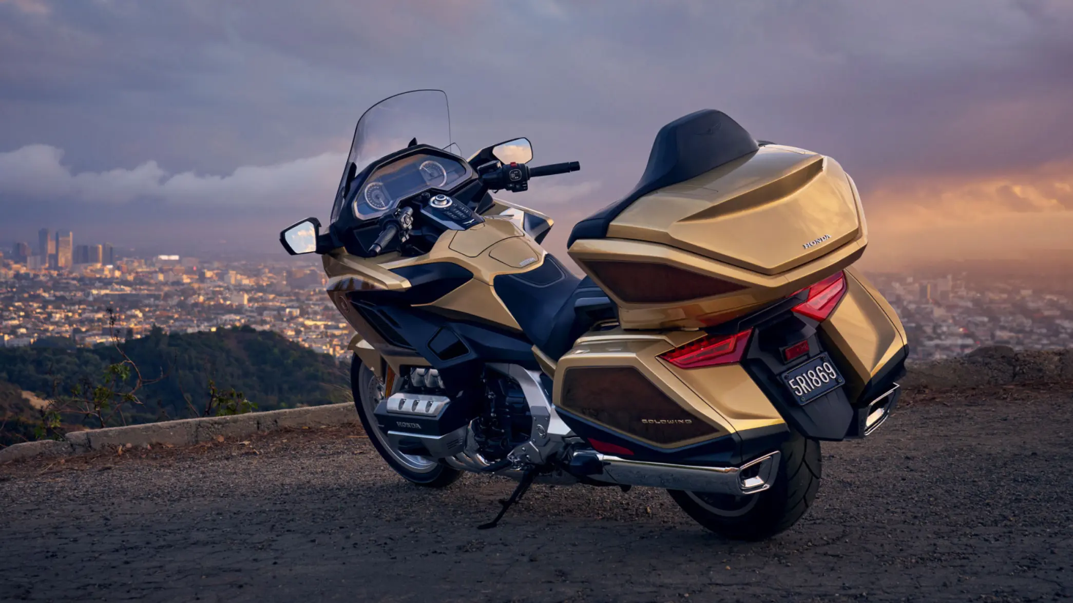 Ein goldenes Honda Goldwing Motorrad steht auf einem Hügel mit Blick auf eine Stadtlandschaft im Hintergrund.