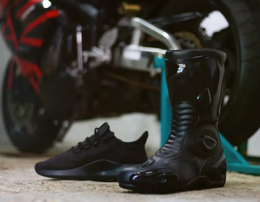 Ein schwarzer Motorradstiefel und ein schwarzer Sneaker stehen nebeneinander vor einem unscharfen Motorrad im Hintergrund.