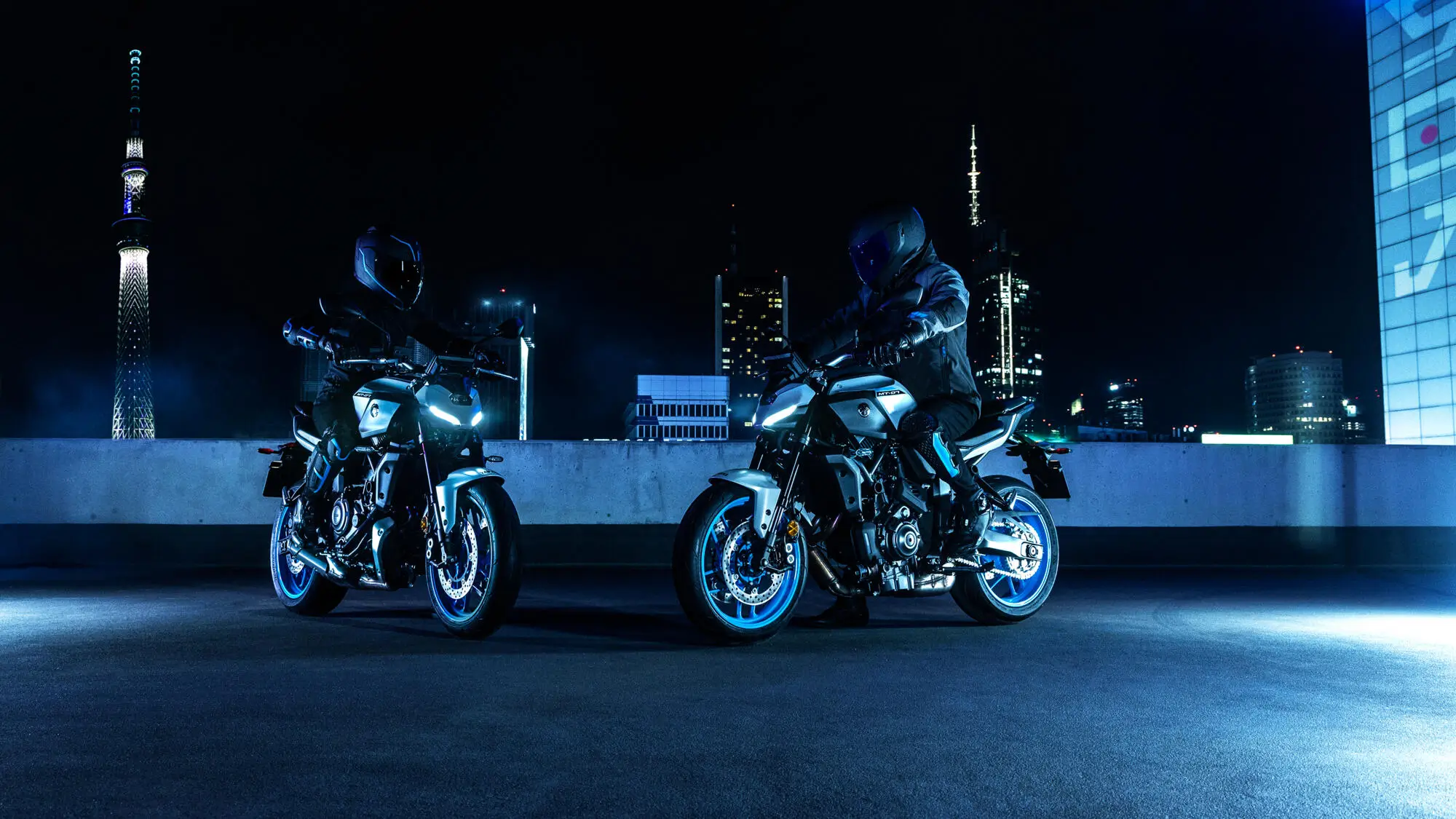 2025-Yamaha-MT07AS-EU-Ice_Storm-Static-001-03 Zwei Personen auf Yamaha MT-07 Motorrädern bei Nacht in einer städtischen Umgebung.