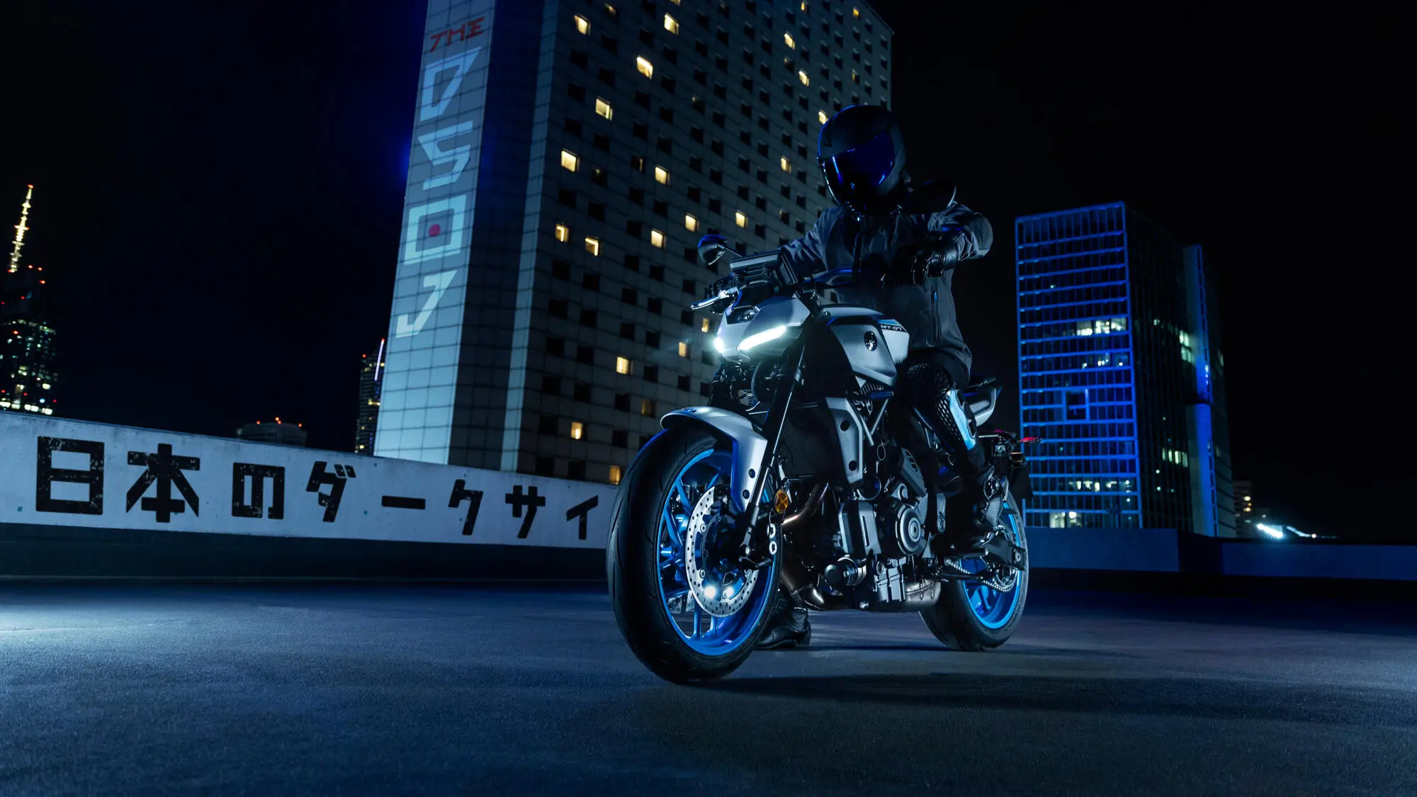 2025-Yamaha-MT07AS-EU-Ice_Storm-Static-002-03 Ein Motorradfahrer auf einer Yamaha MT-07 AS bei Nacht in einer städtischen Umgebung.