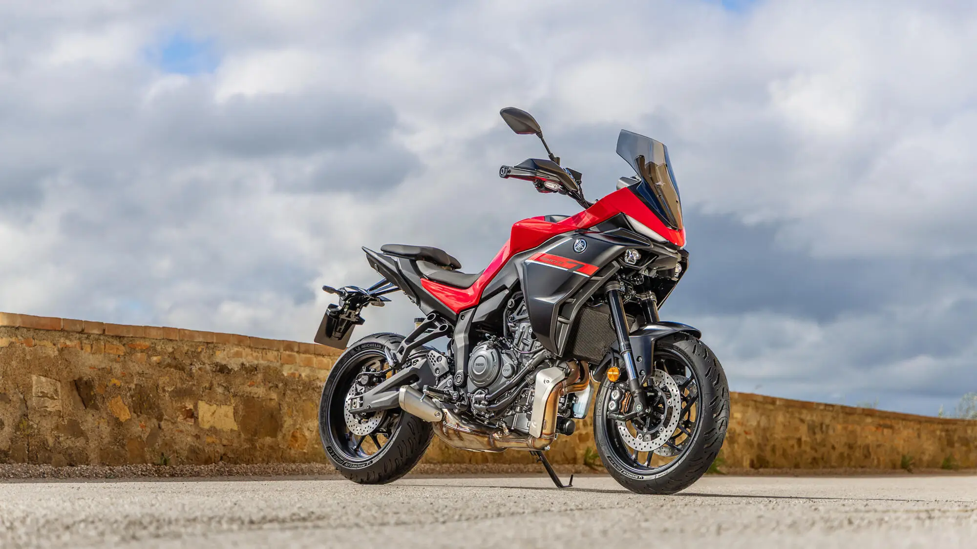 2025-Yamaha-MT07TR-EU-Redline-Static-001-03 Ein rotes Yamaha MT-07 TR Motorrad steht auf einer Straße vor einer Steinmauer.