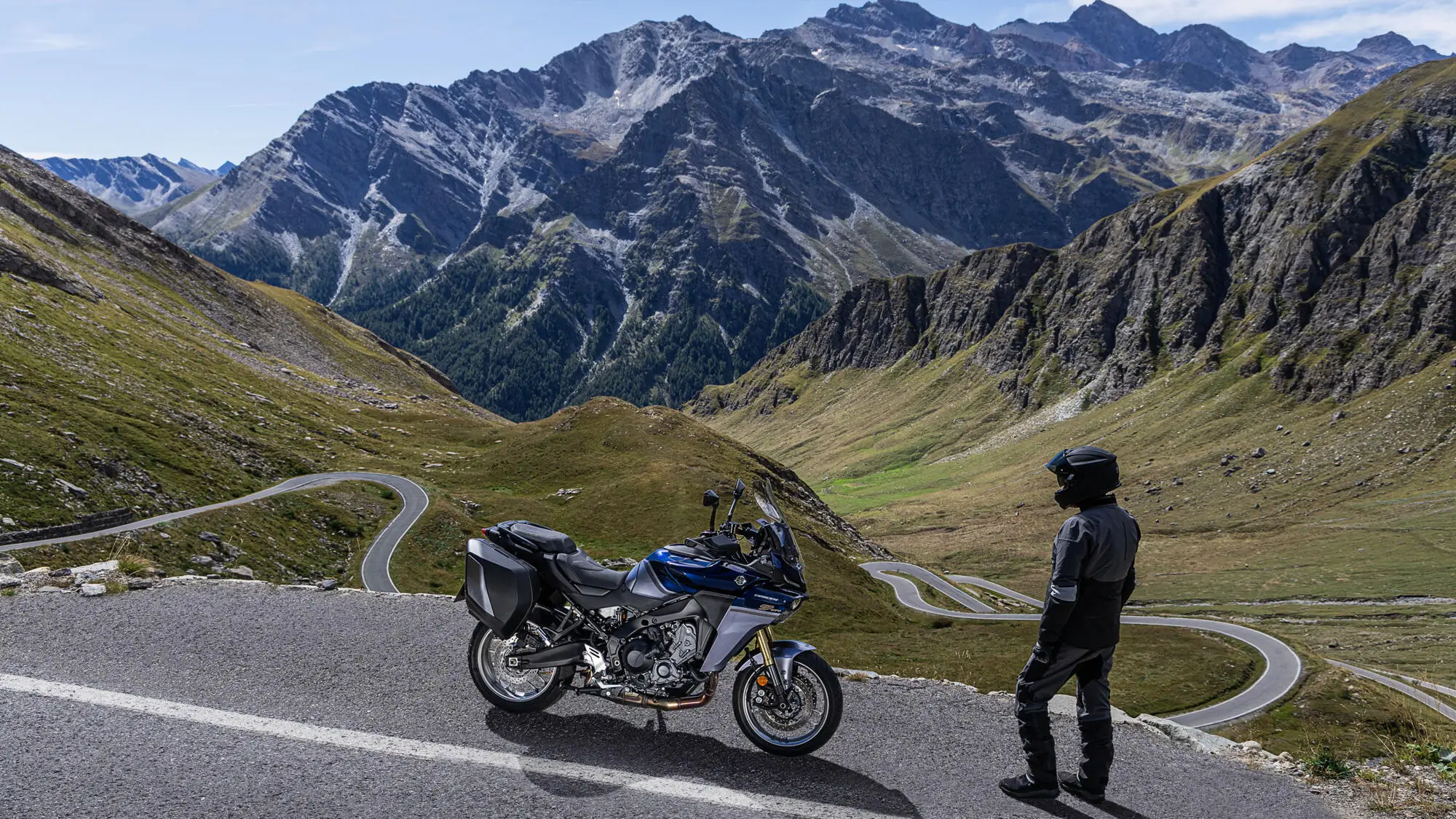 Ein Motorradfahrer steht neben einer Yamaha TRACER 9 GT+ auf einer kurvigen Bergstraße mit beeindruckender Bergkulisse.