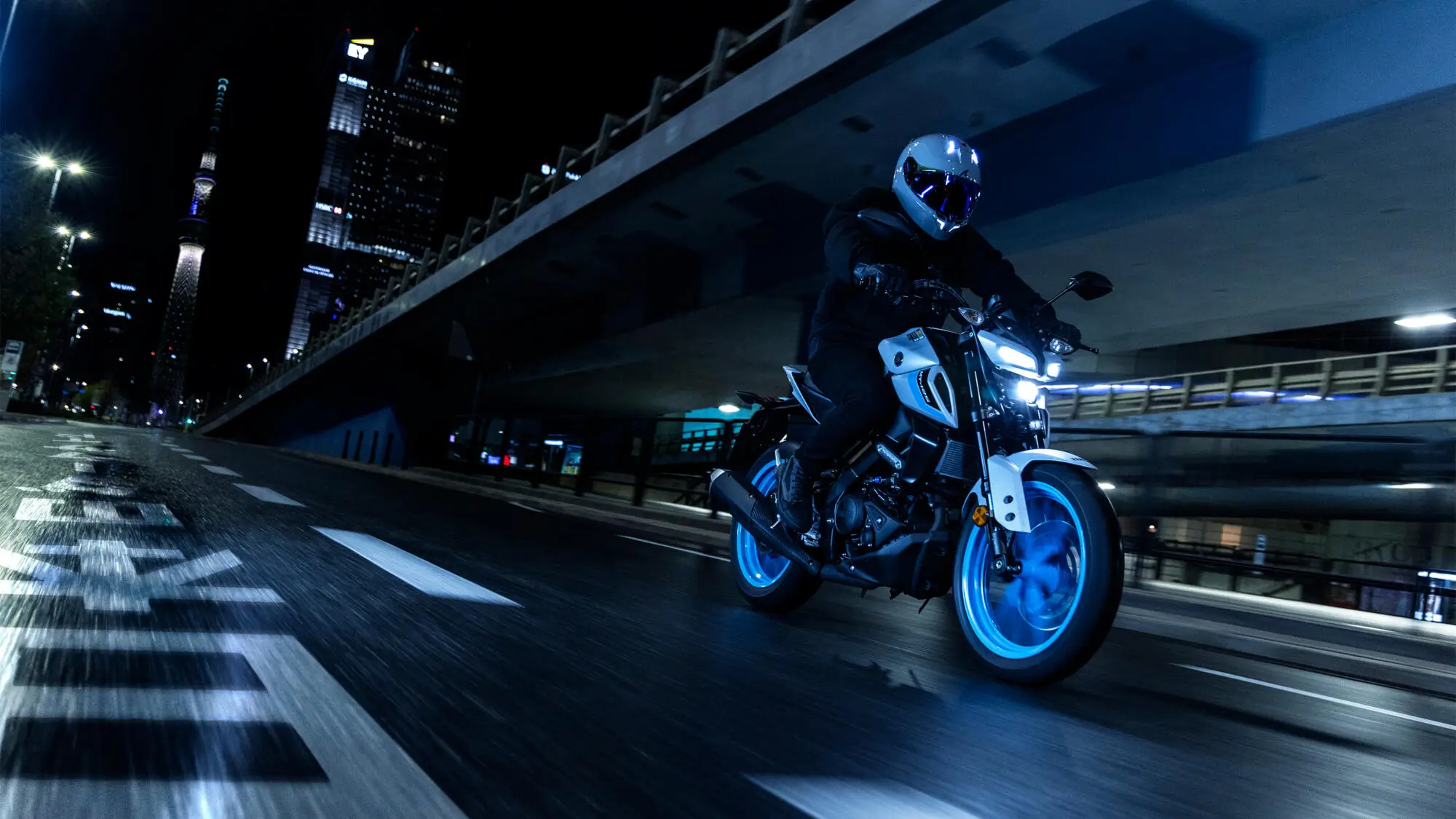 2025-Yamaha-MT125A-EU-Ice_Storm-Action-001-03 Ein Motorradfahrer fährt nachts auf einer Yamaha MT-125 durch eine beleuchtete Stadt.