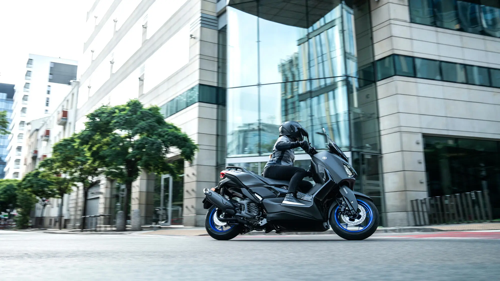 Eine Person fährt auf einem Yamaha XMAX 125A Roller durch eine städtische Umgebung.