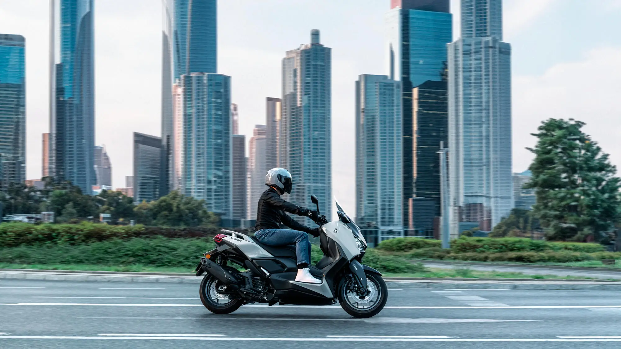 Eine Person fährt einen Yamaha XMAX 125 Roller vor einer Skyline mit Wolkenkratzern.