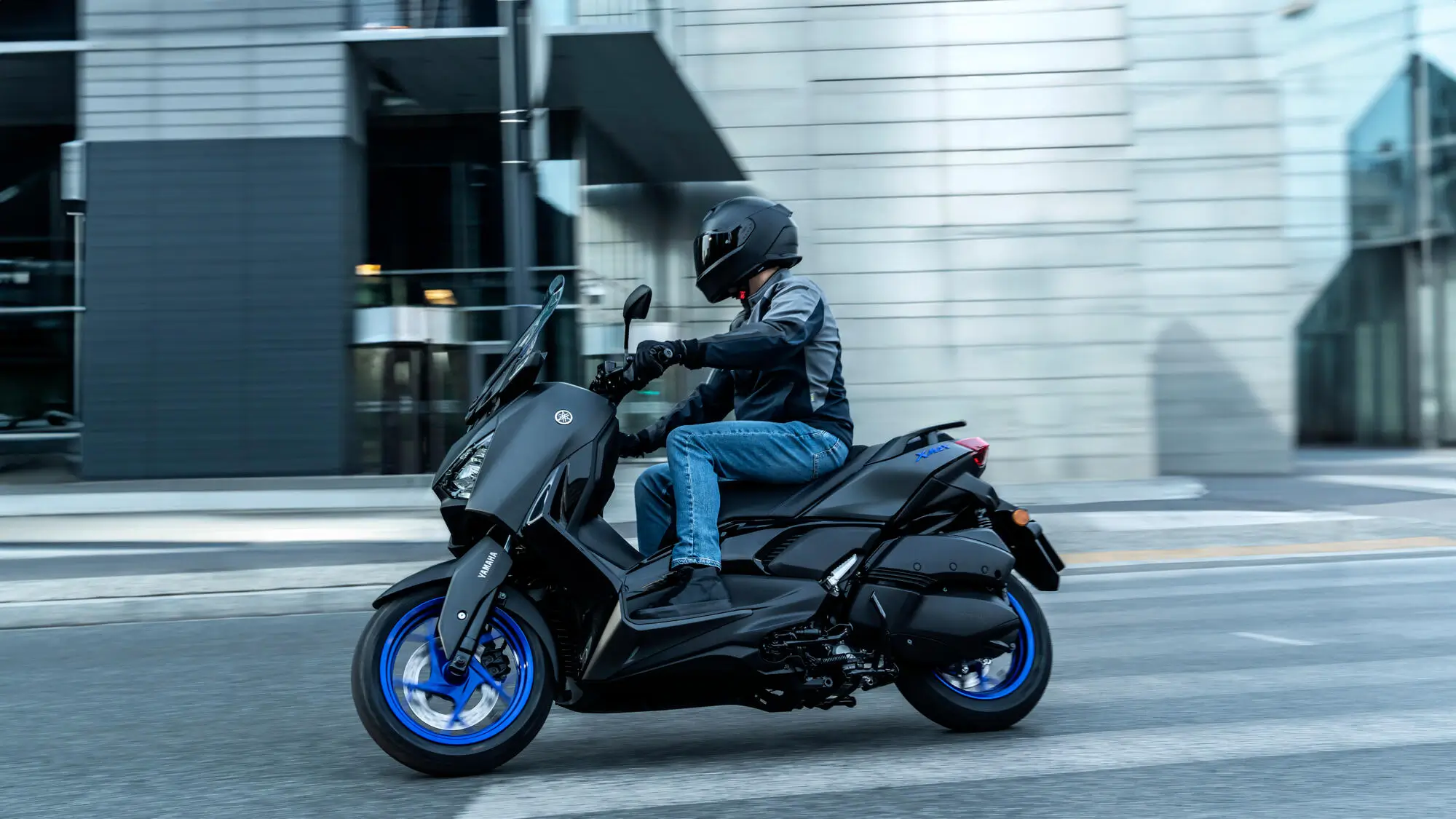 Ein Fahrer auf einem schwarzen Yamaha XMAX 300A Roller fährt durch eine städtische Umgebung.