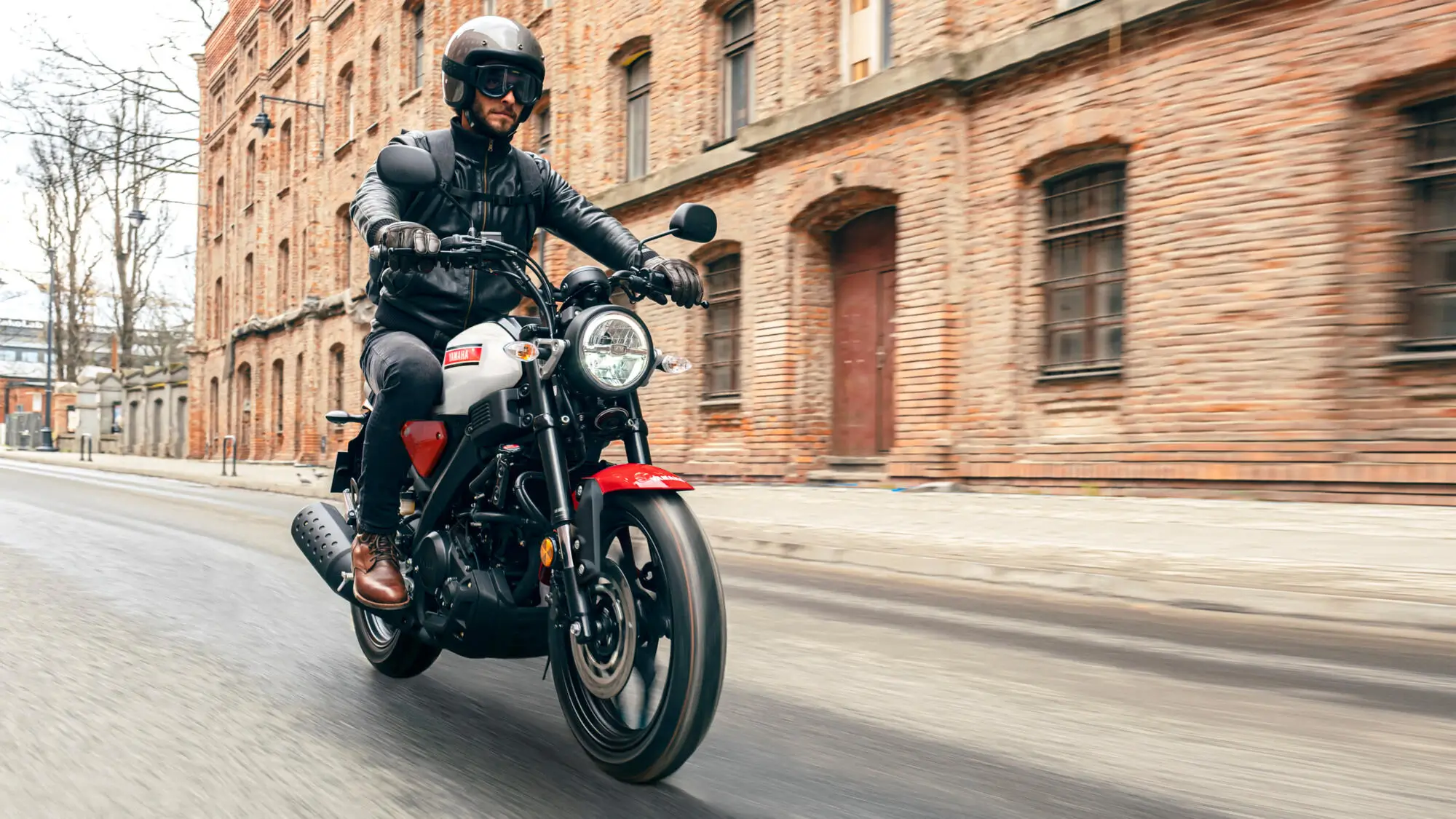 2025-Yamaha-XS125-EU-Legend_White-Action-004-03 Ein Fahrer auf einer Yamaha XS125 fährt an einem Backsteingebäude vorbei.