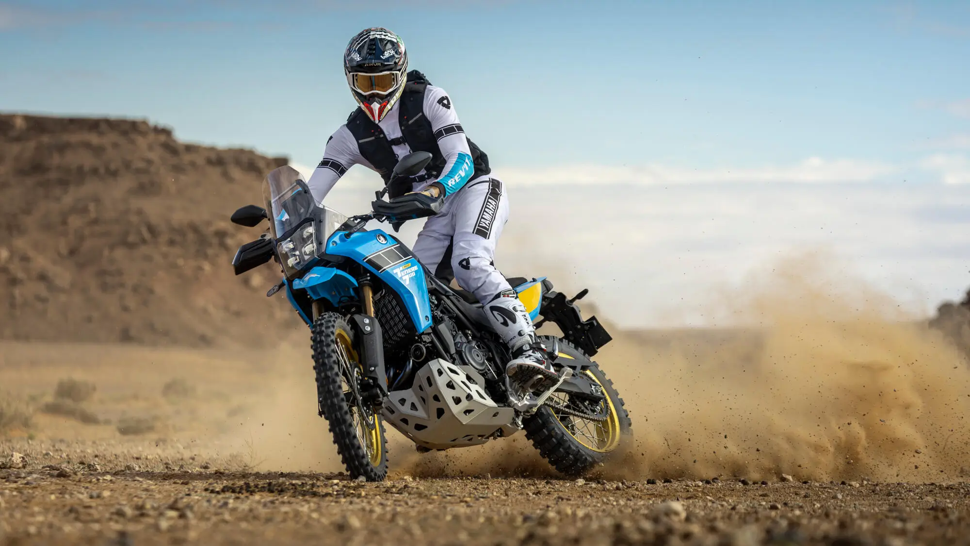 2025-Yamaha-XTZ700SPR-EU-Sky_Blue-Action-016-03 Ein Motorradfahrer fährt eine Yamaha XTZ700SPR im Gelände und wirbelt Staub auf.