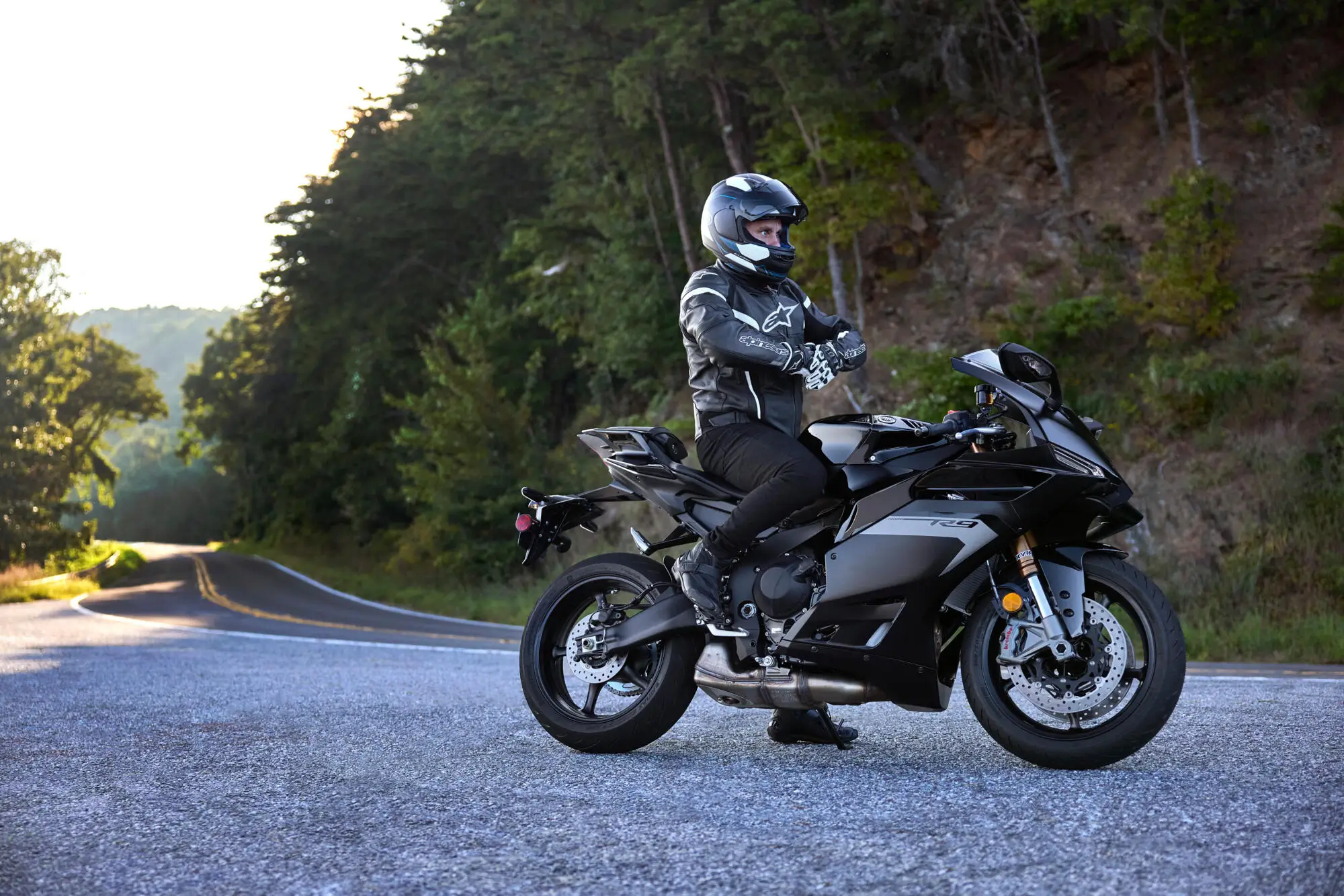 Ein Motorradfahrer auf einer Yamaha YZF900R9 steht auf einer kurvigen Straße in einer waldreichen Umgebung.