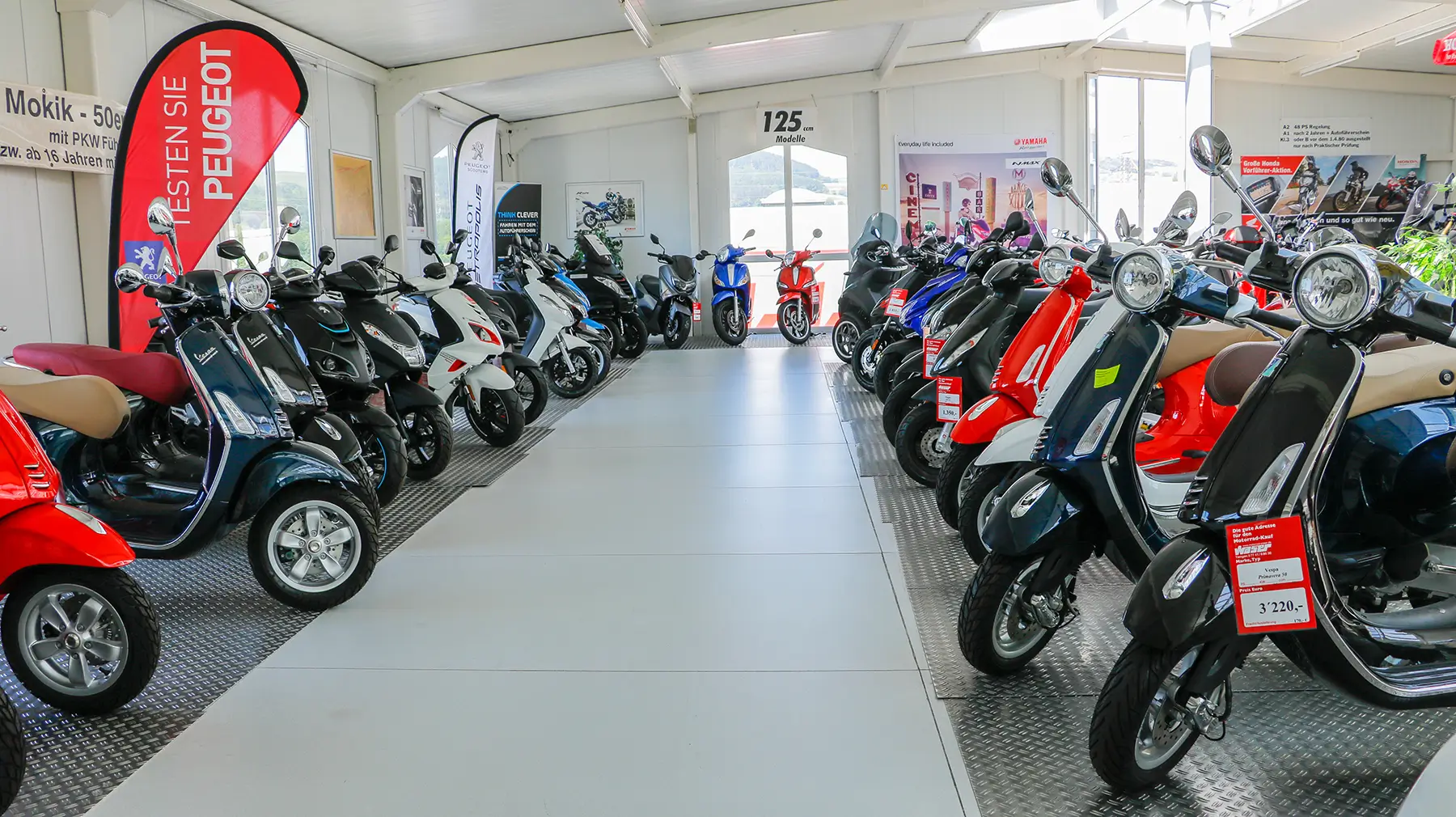 Eine Ausstellungshalle voller verschiedener Motorroller, darunter Modelle von Peugeot.