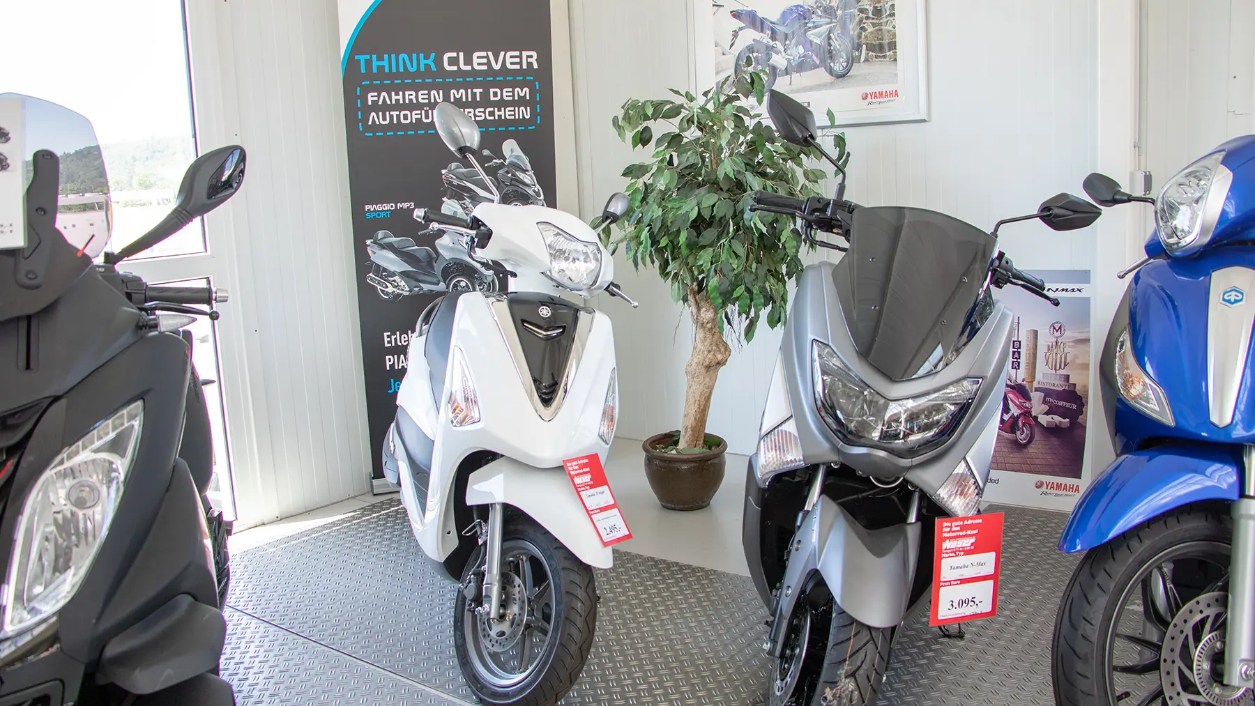 Drei Motorroller, darunter ein Yamaha NMAX, stehen in einer Ausstellungshalle.