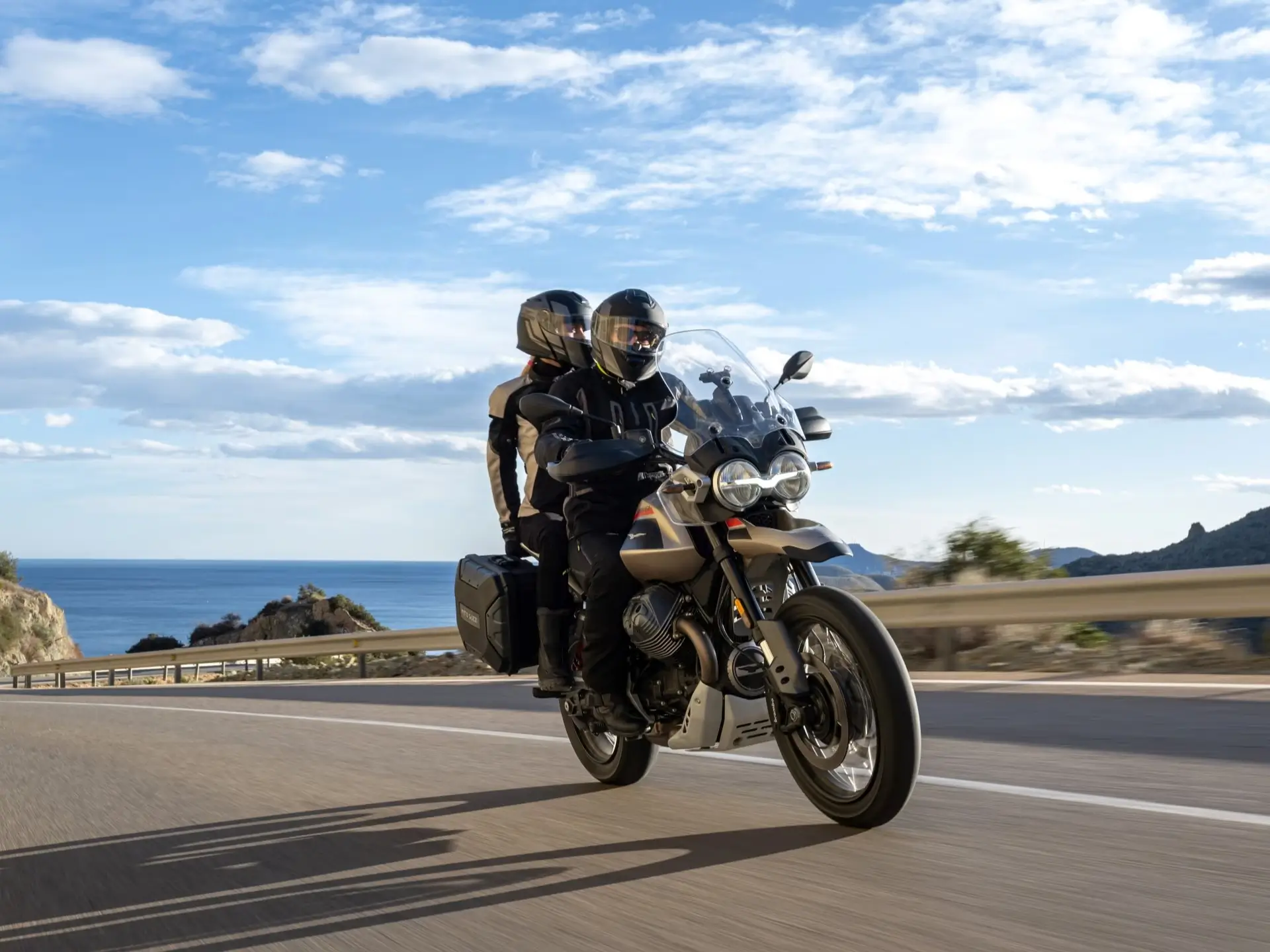 Moto Guzzi V85 TT Travel mit Beifahrer auf Küstenstraße, Gepäckkoffer.
