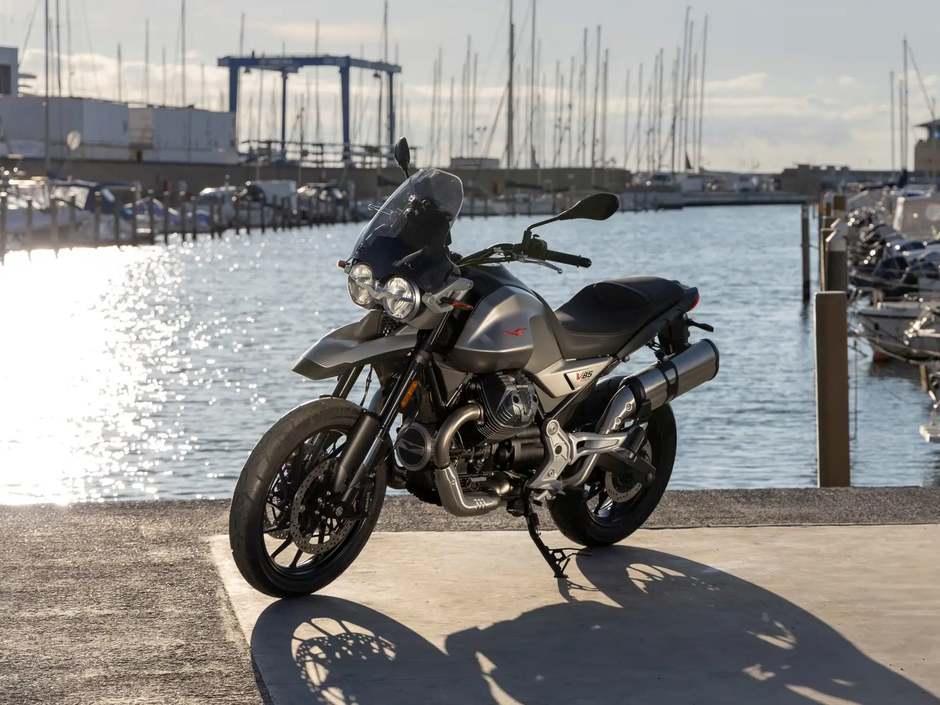 Moto Guzzi V85 Strada steht am Hafen, Boote im Hintergrund.
