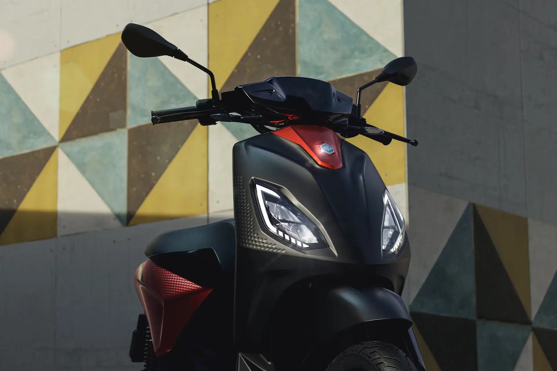Piaggio-Scooter, schwarzer Frontkorpus mit rotem Akzent, geometrische Wand im Hintergrund.