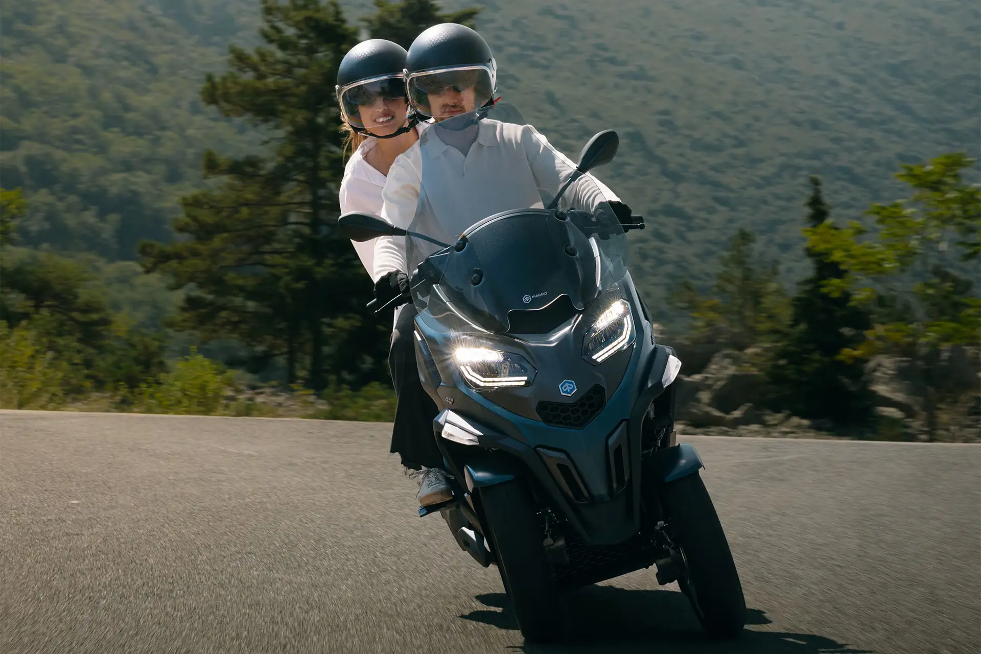 PiaggioMP3-530-gallery2 Zwei Personen fahren auf einem Piaggio MP3 530 Roller auf einer Straße in einer grünen Landschaft.