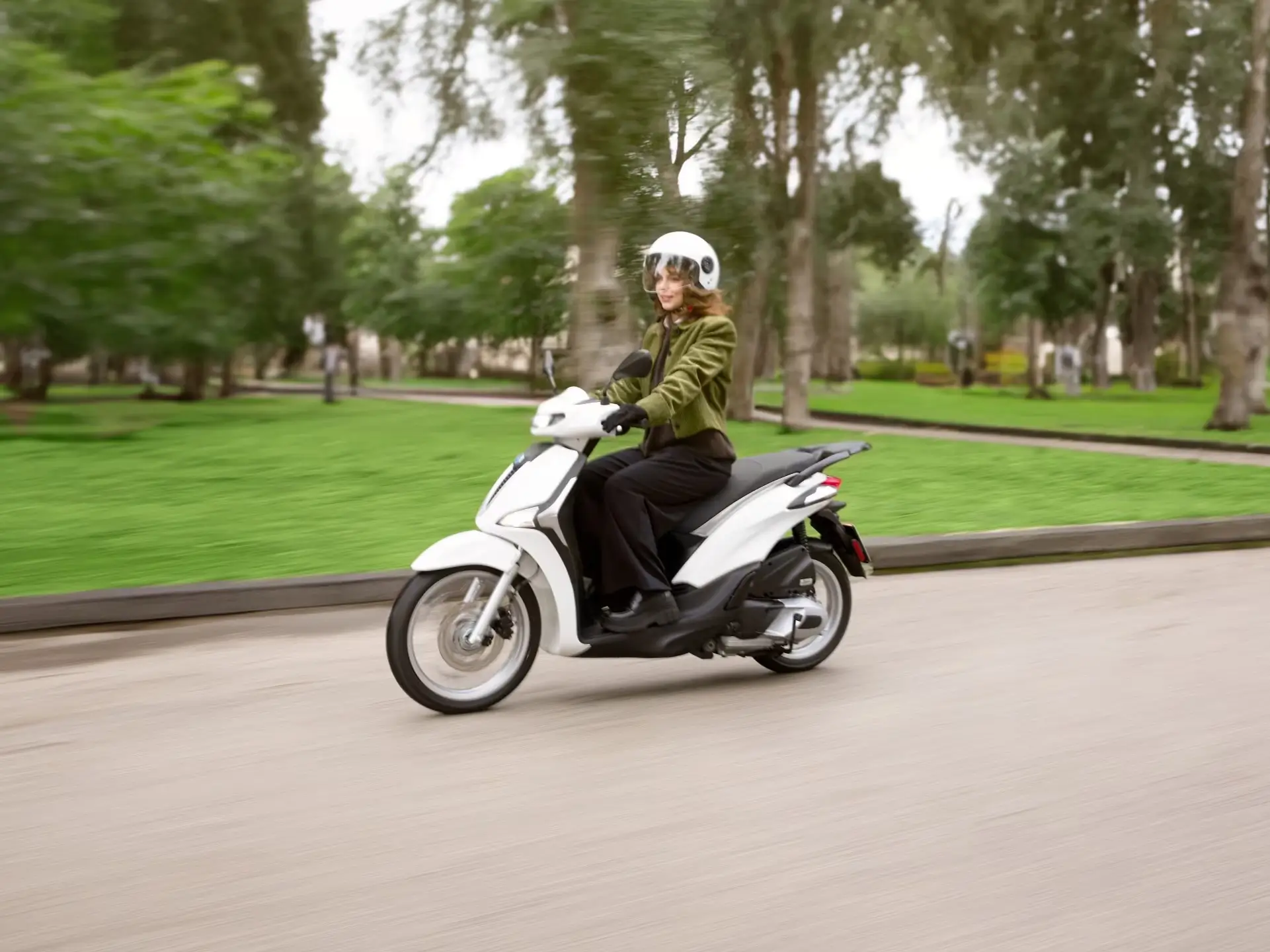 Eine Person fährt auf einem weißen Piaggio Liberty Roller durch einen Park.