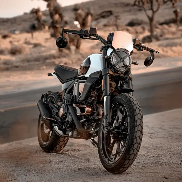 Scrambler-Ducati-10°-Anniversario-Rizoma-Edition-MY25-flexicard-1080x1080-01-1-768x768 Eine Ducati Scrambler 10° Anniversario Rizoma Edition steht in einer wüstenähnlichen Landschaft.