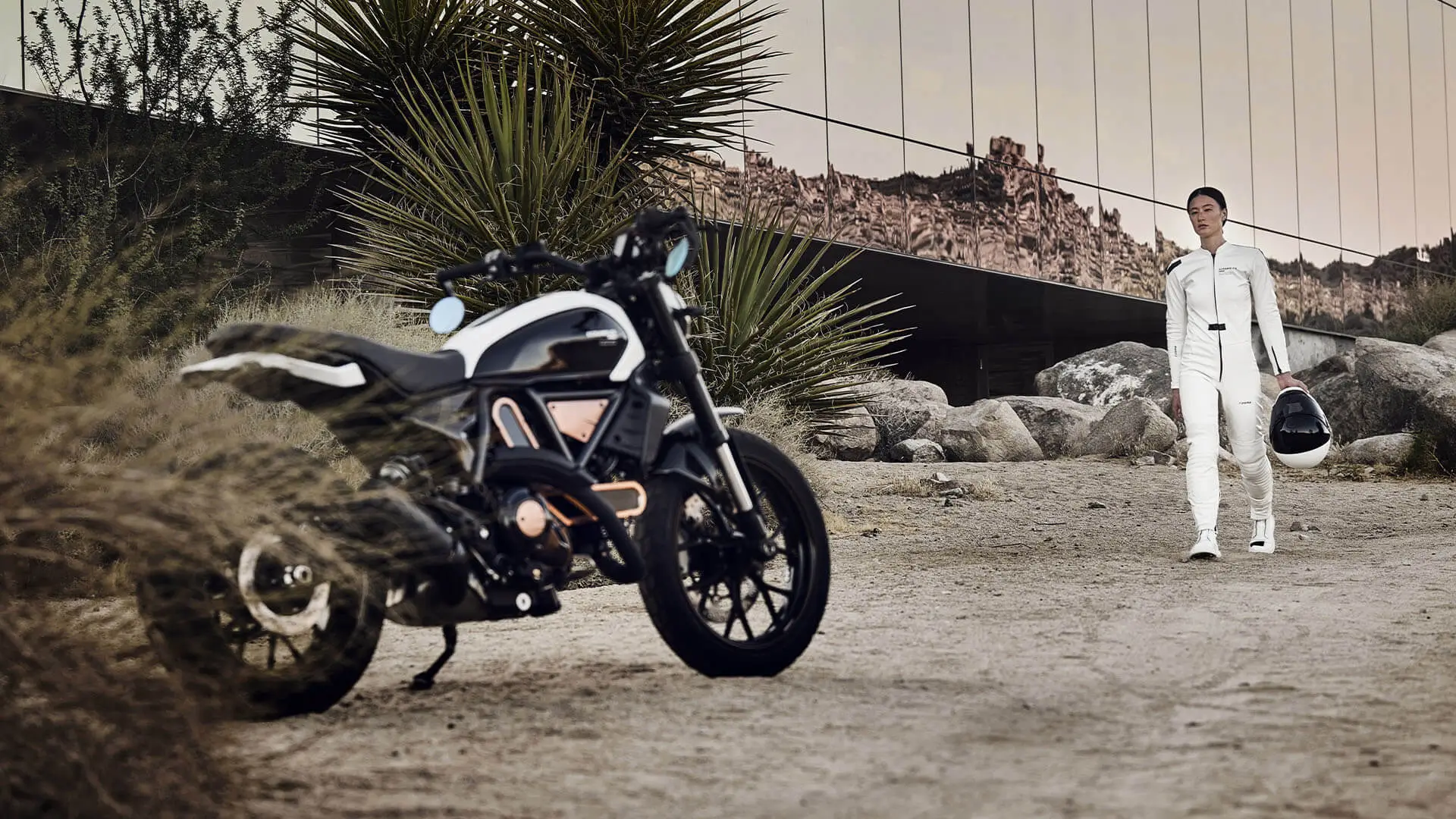 Scrambler-Ducati-10°-Anniversario-Rizoma-Edition-MY25-media-slider-02-1920x1080-01 Eine Person in weißem Anzug trägt einen Helm und geht auf ein Ducati Scrambler Motorrad zu.