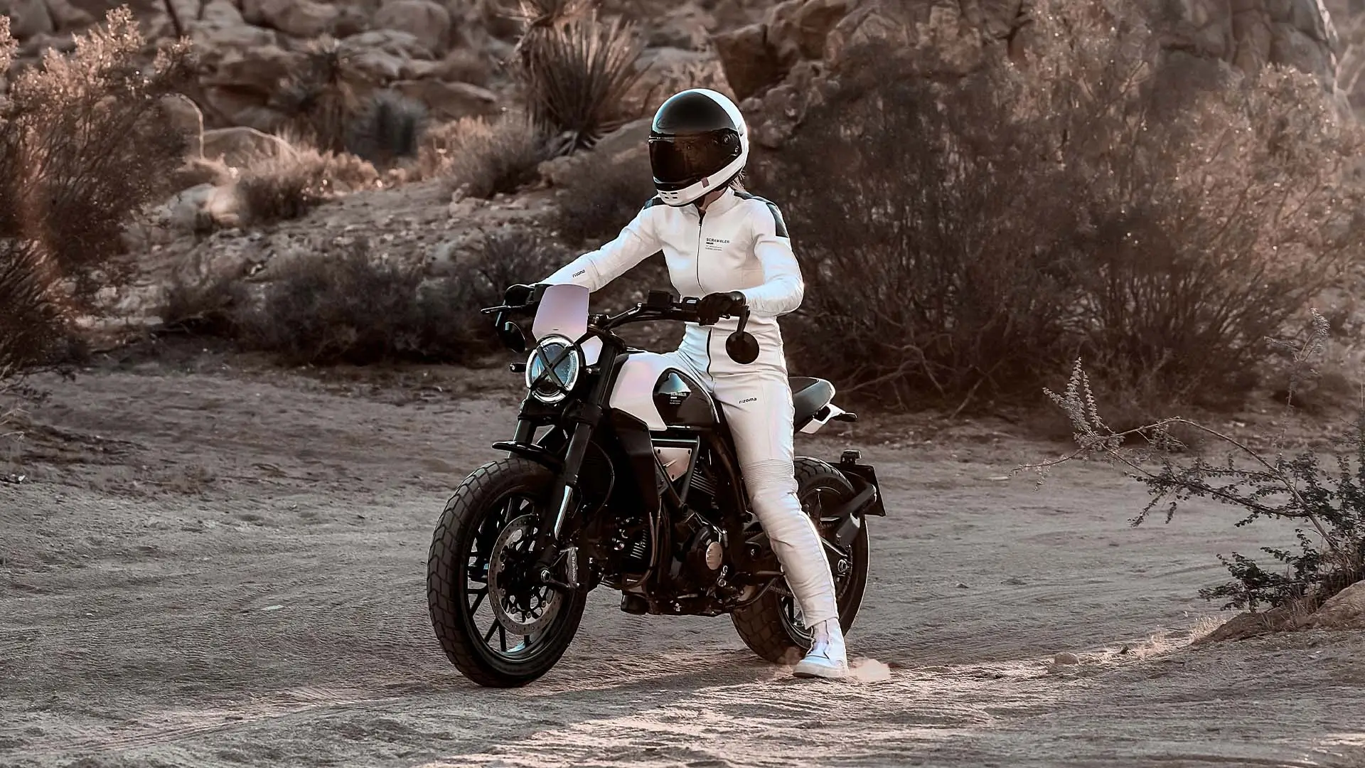 Scrambler-Ducati-10°-Anniversario-Rizoma-Edition-MY25-media-slider-02-1920x1080-08 Eine Person in weißer Motorradkleidung fährt eine Ducati Scrambler in einer Wüstenlandschaft.