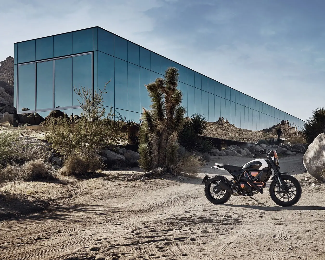 Ein Ducati Scrambler Motorrad steht vor einem modernen, spiegelnden Gebäude in einer Wüstenlandschaft.