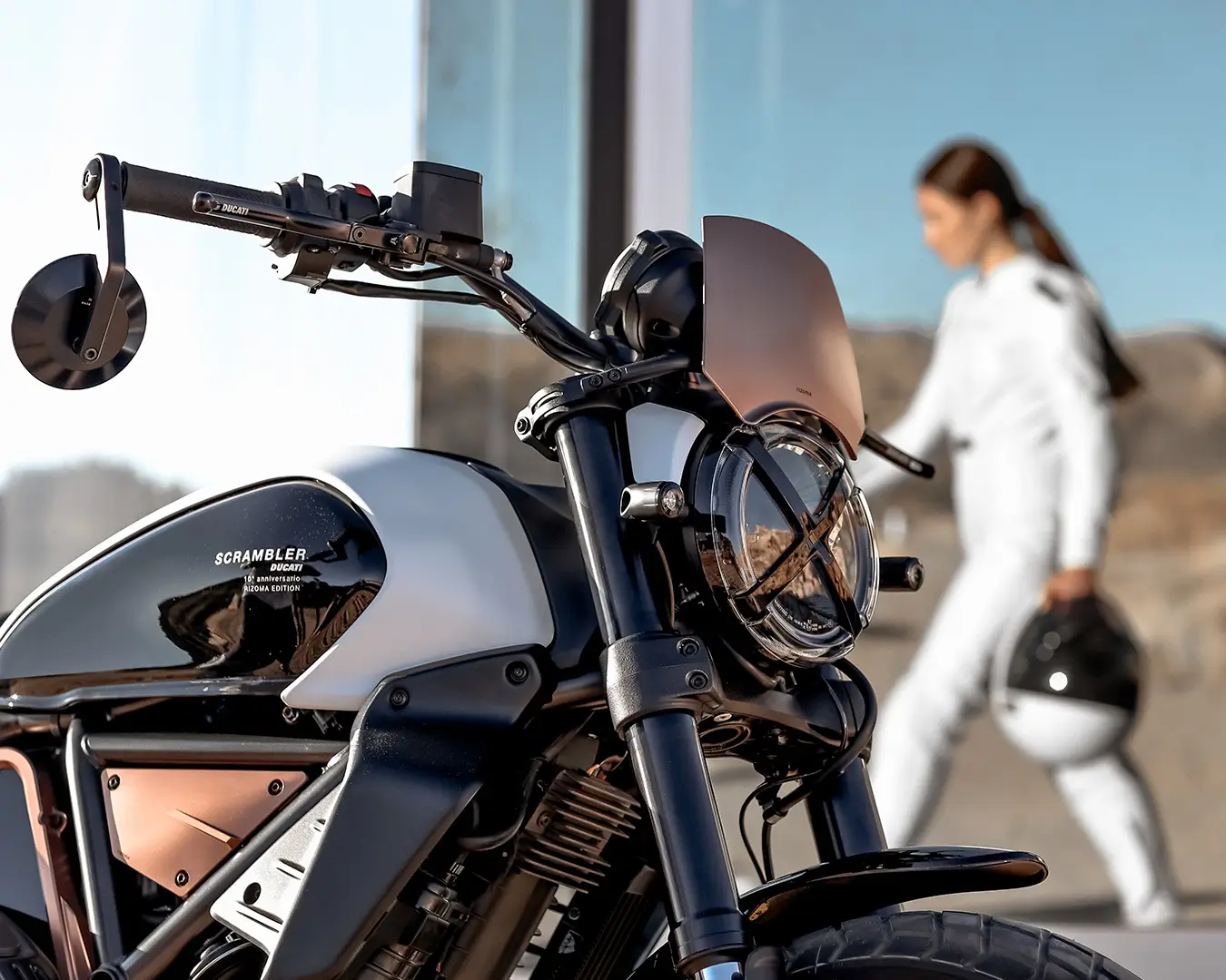 Ducati Scrambler 10° Anniversario Rizoma Edition im Vordergrund, Person in Motorradkleidung im Hintergrund.