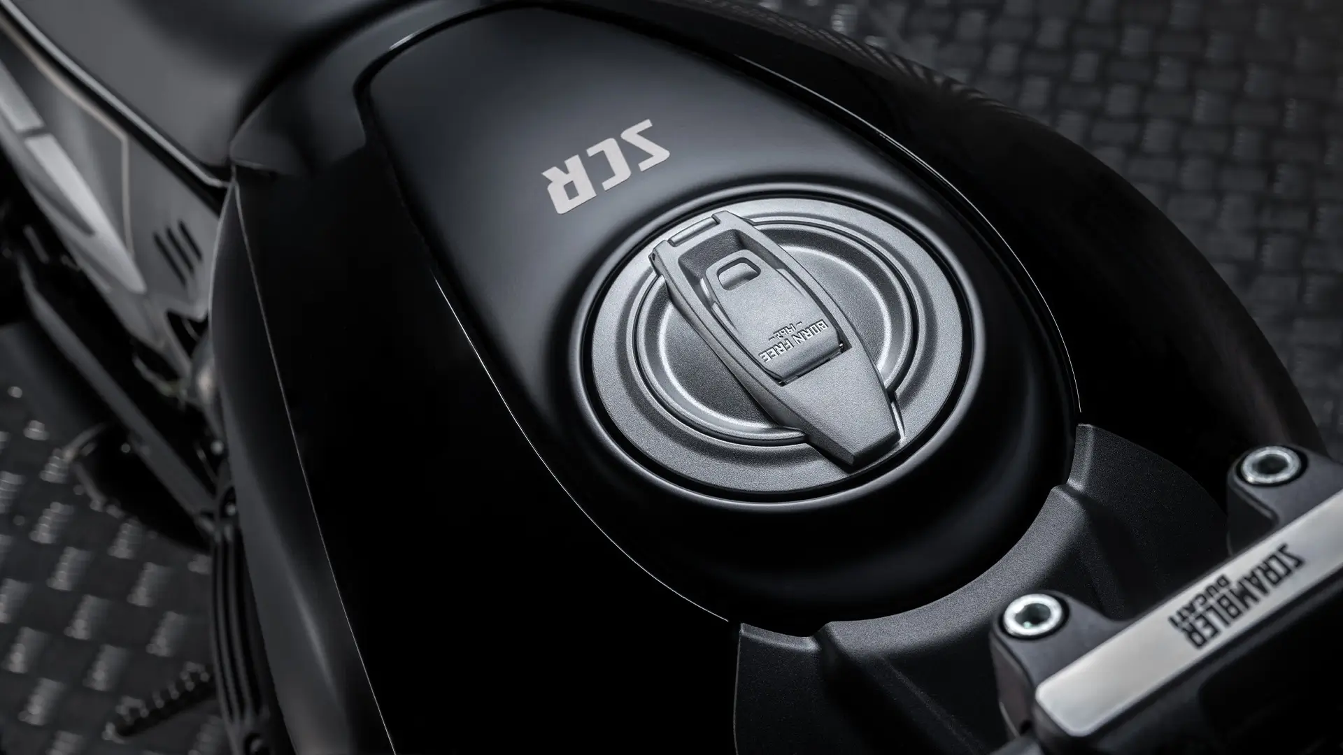 Nahaufnahme eines Motorradtanks mit der Aufschrift "SCR" und einem Tankdeckel.