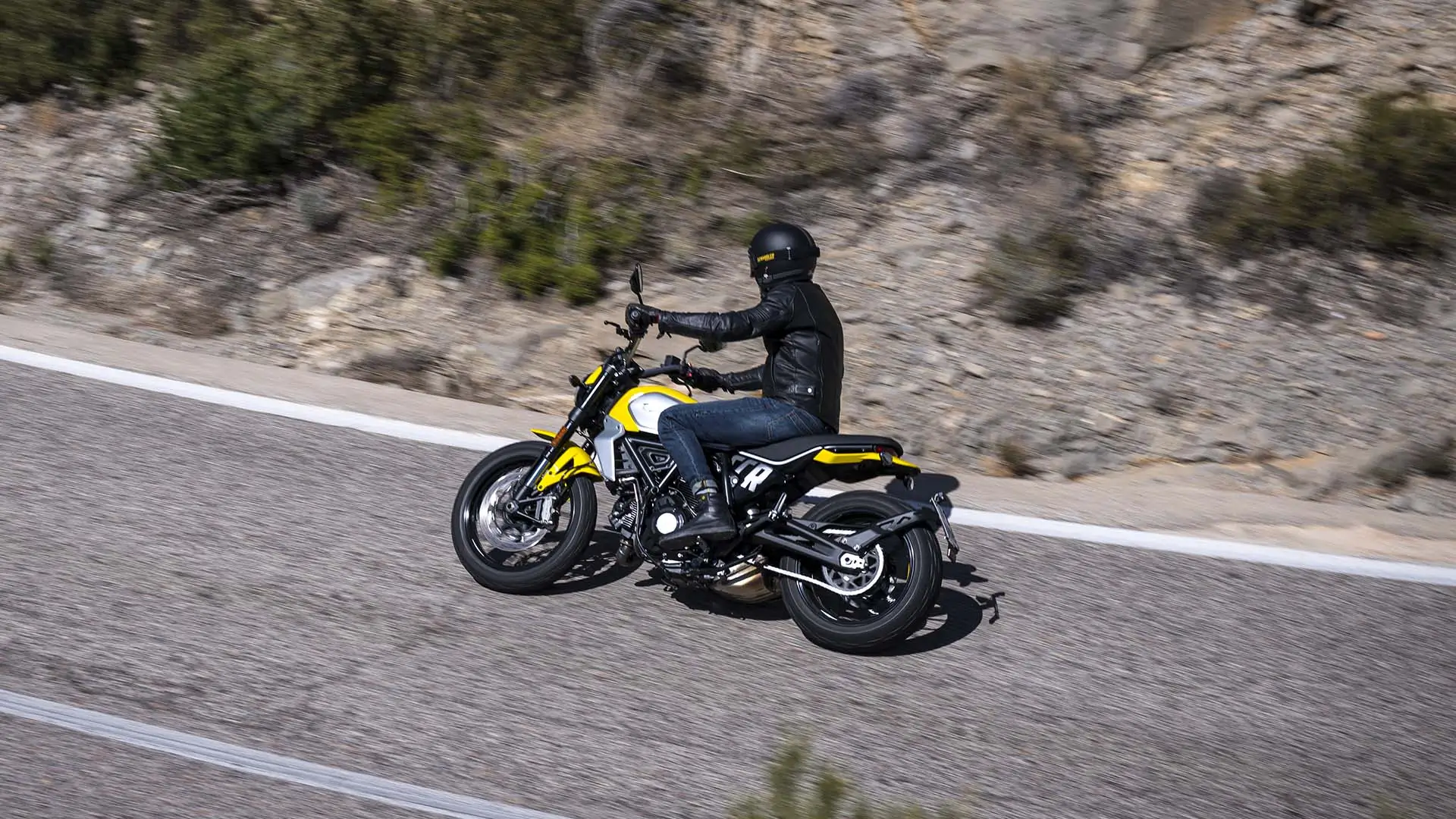 Ein Motorradfahrer auf einer gelben Scrambler Icon fährt auf einer Straße entlang.