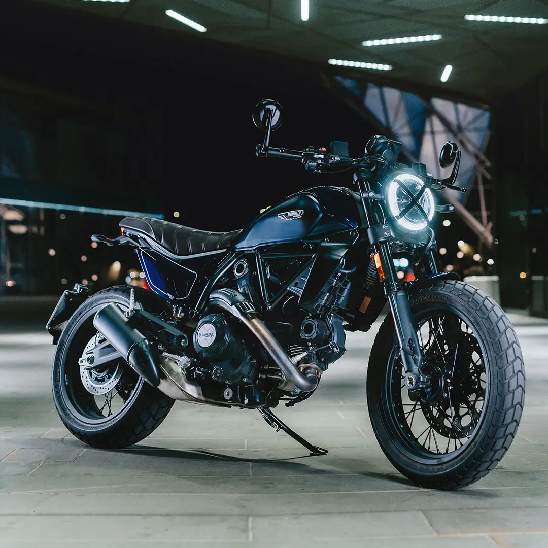 Scrambler-single-bike-nightshift-mosaic-media-2-1080x1080-03 Ein Scrambler-Motorrad in einer nächtlichen, urbanen Umgebung.