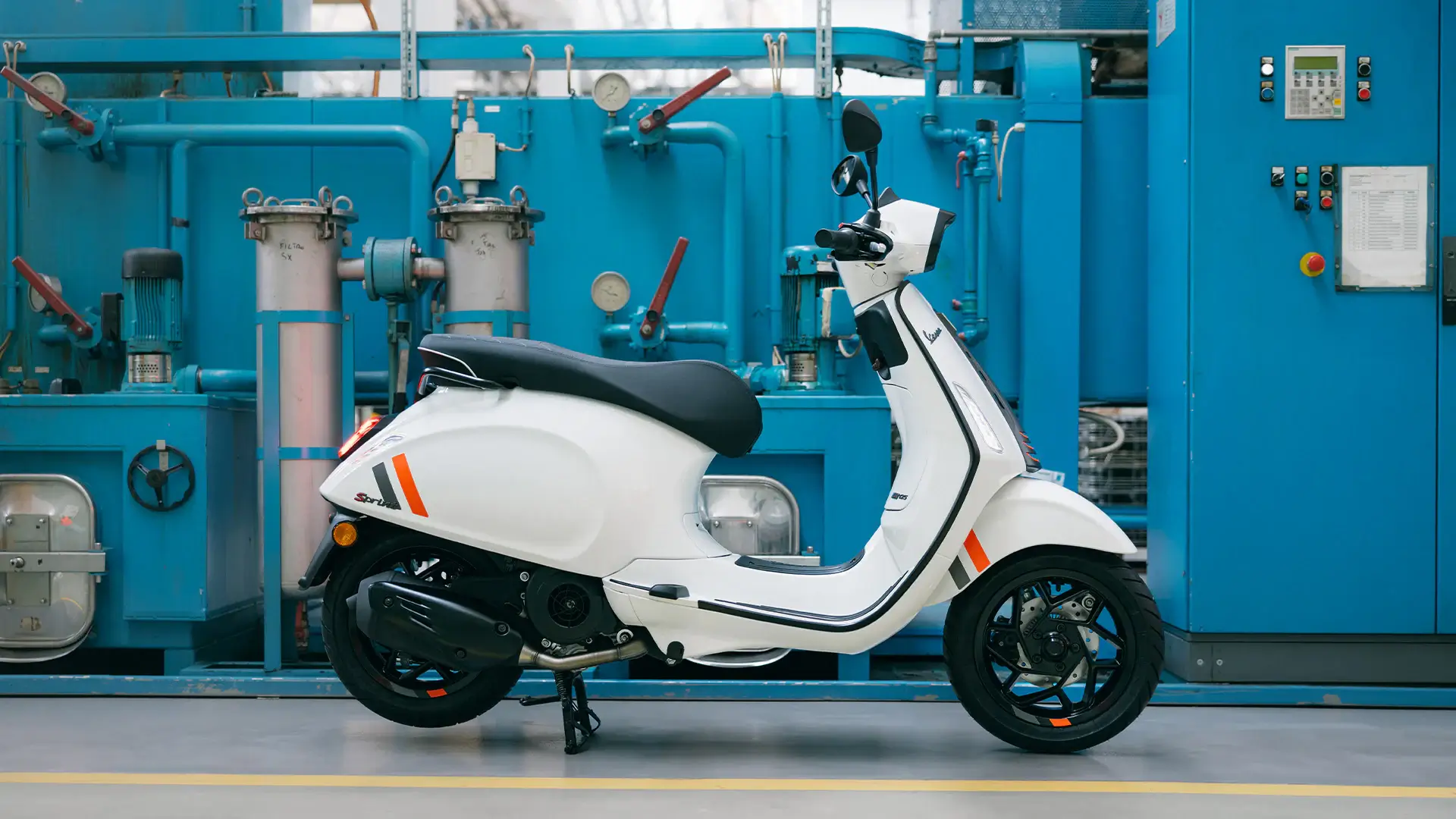 Eine weiße Vespa Sprint steht vor einer blauen industriellen Anlage.