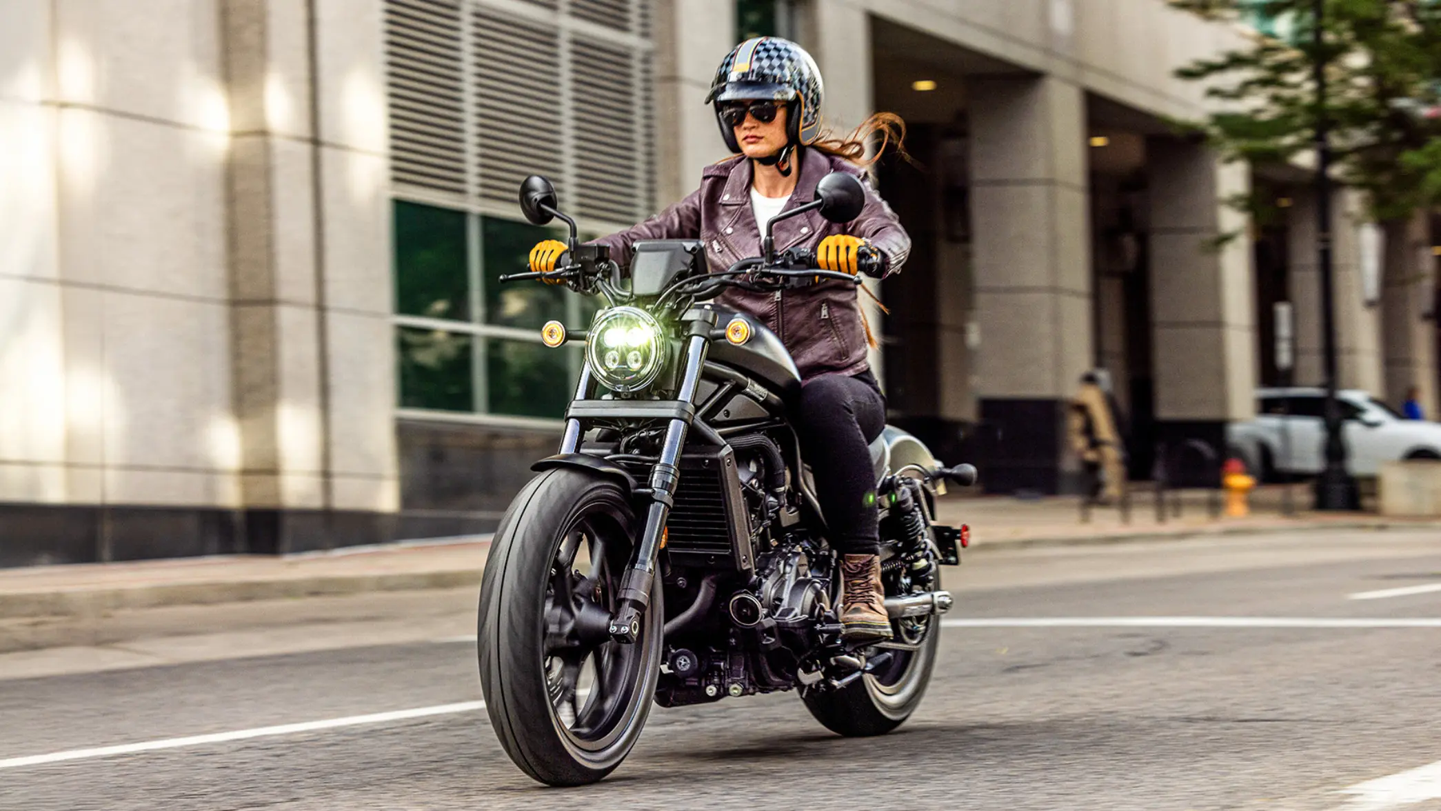 Frau fährt ein Motorrad Honda CMX1100 Rebel in der Stadt, trägt Helm und Sonnenbrille.