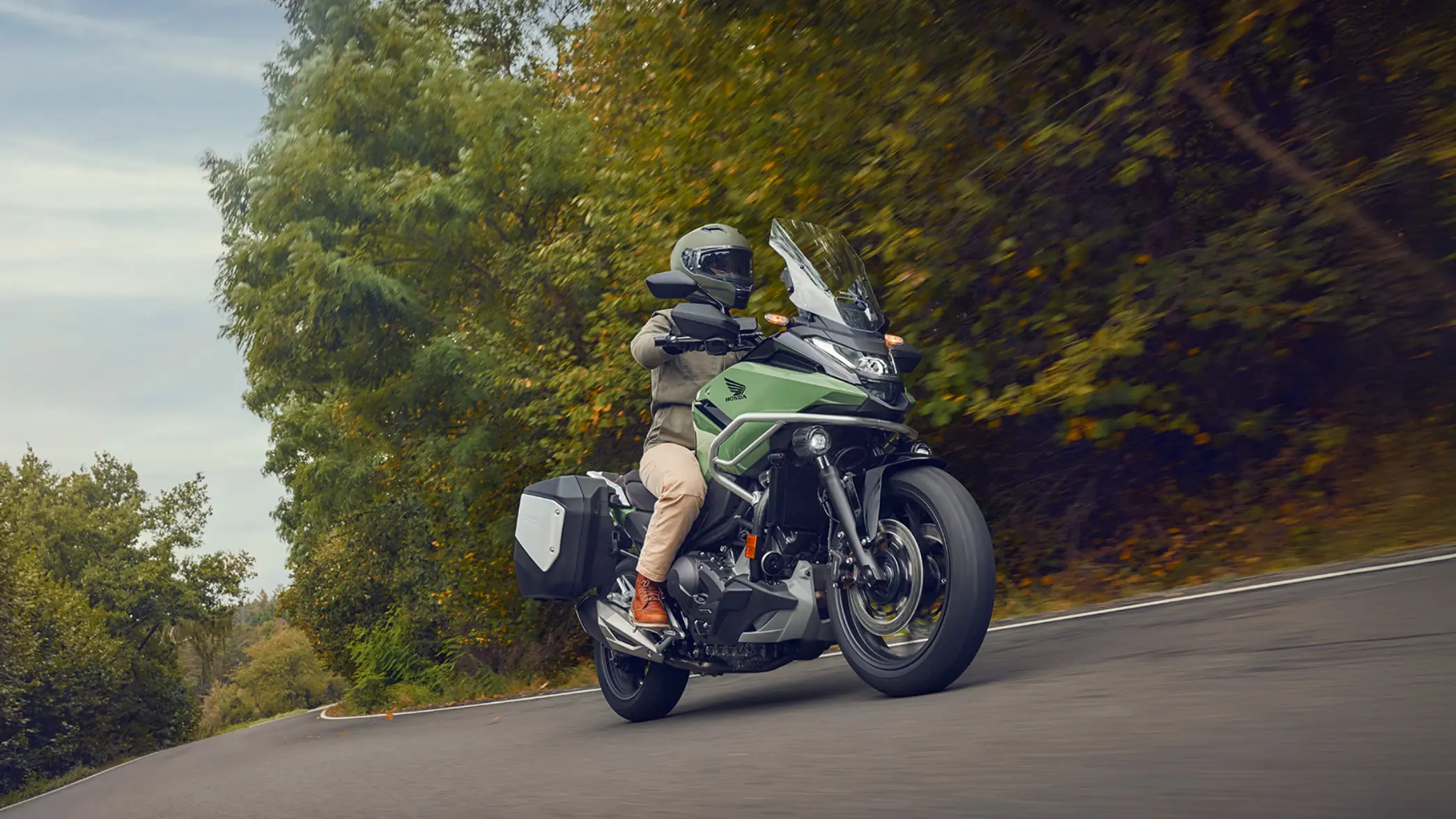 Ein Motorradfahrer auf einer grünen Honda NC750X fährt auf einer kurvigen Straße durch eine bewaldete Landschaft.