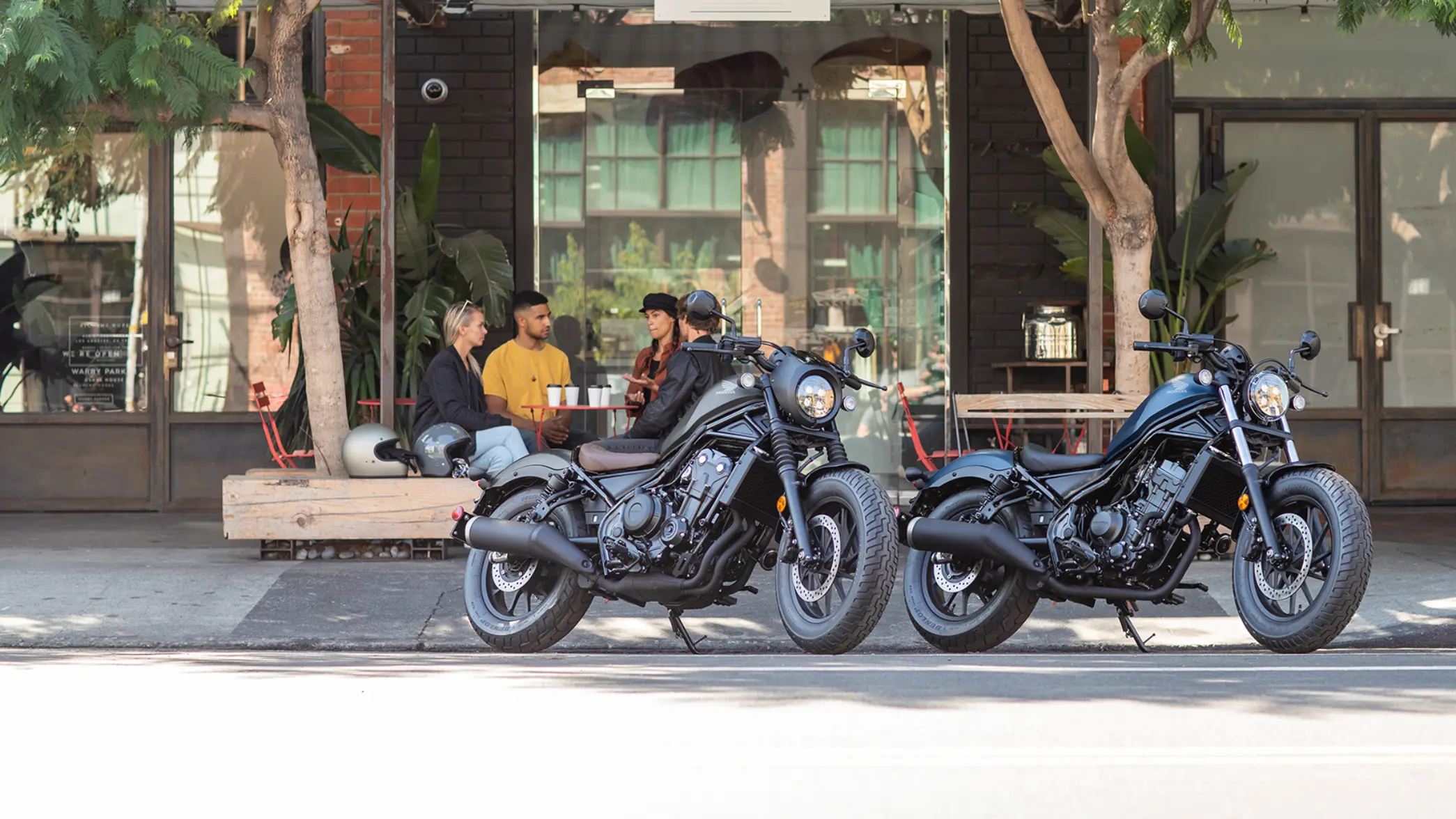 fb_r_w Zwei Honda CMX500 REBEL Motorräder parken vor einem Café, während vier Personen an einem Tisch sitzen.